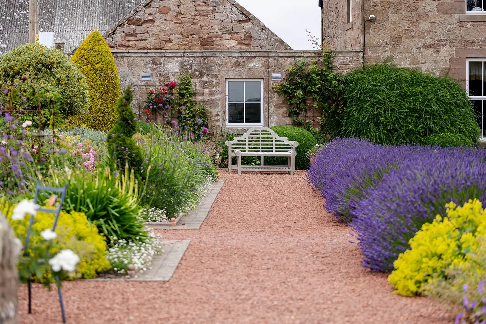 A garden with colorful flowering plants and shrubs lining a gravel pathway, with a white bench against a brick building wall in the background.