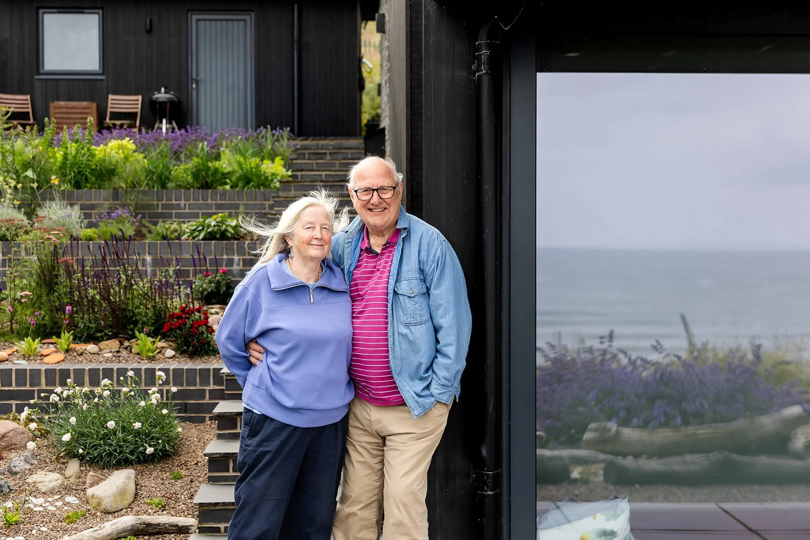 Happy Clients Brian and Rosemary at Sandbank leaning against their home smiling at the camera, the sea is visable in the reflection of the windows