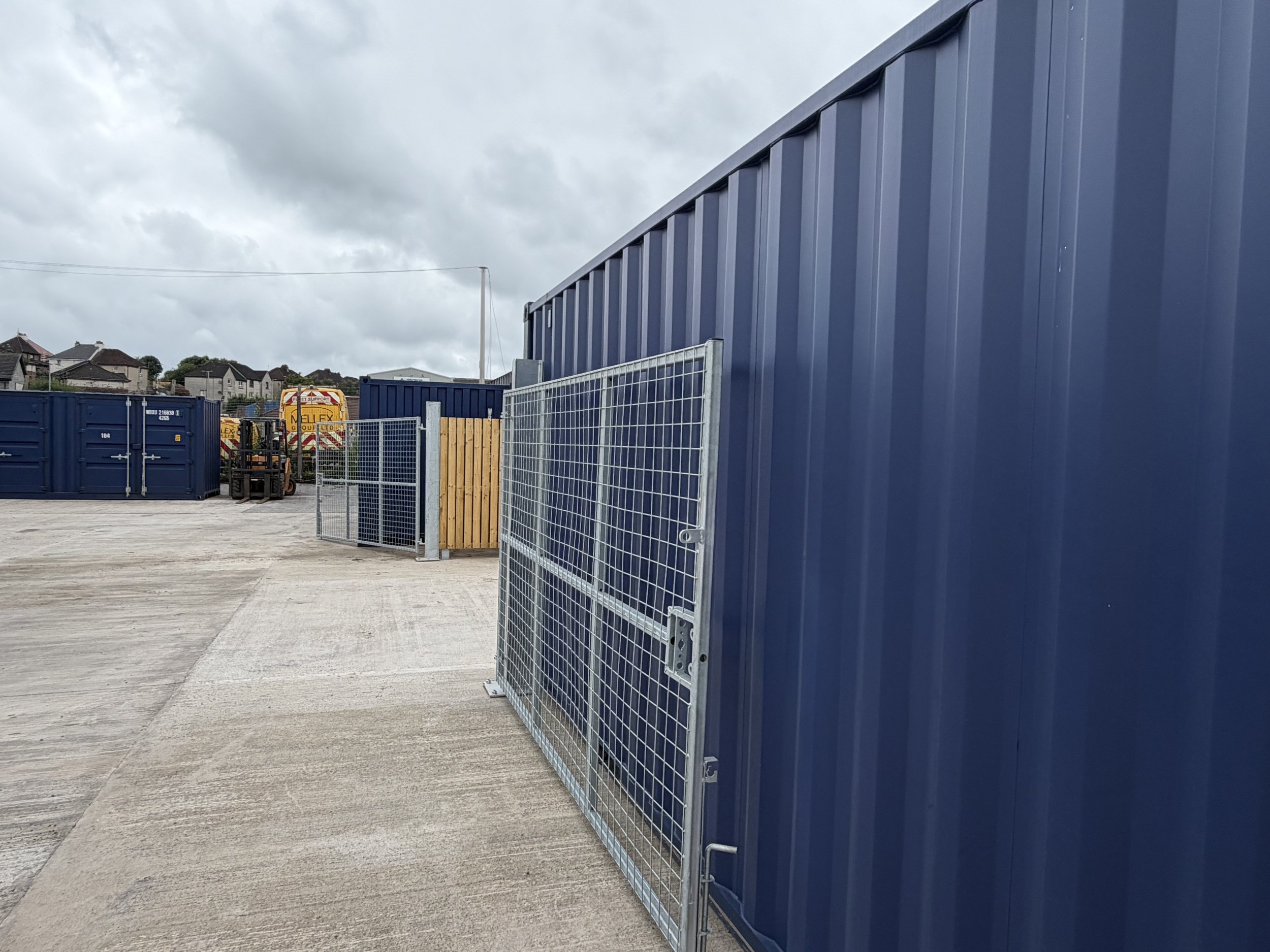 A fenced industrial area with blue storage containers, a wooden gate, a forklift, and a yellow vehicle with a red and white chevron pattern on the back, under a cloudy sky.