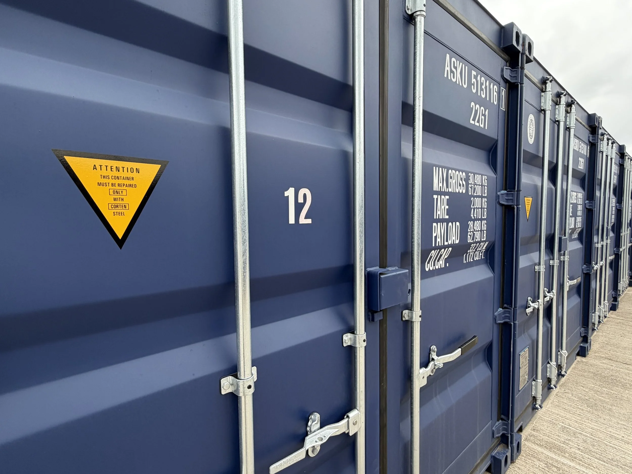 Close-up of blue shipping containers with metal bars and hardware, and a yellow triangular warning sign that says 'Attention: This container must be repaired only with corten steel.'