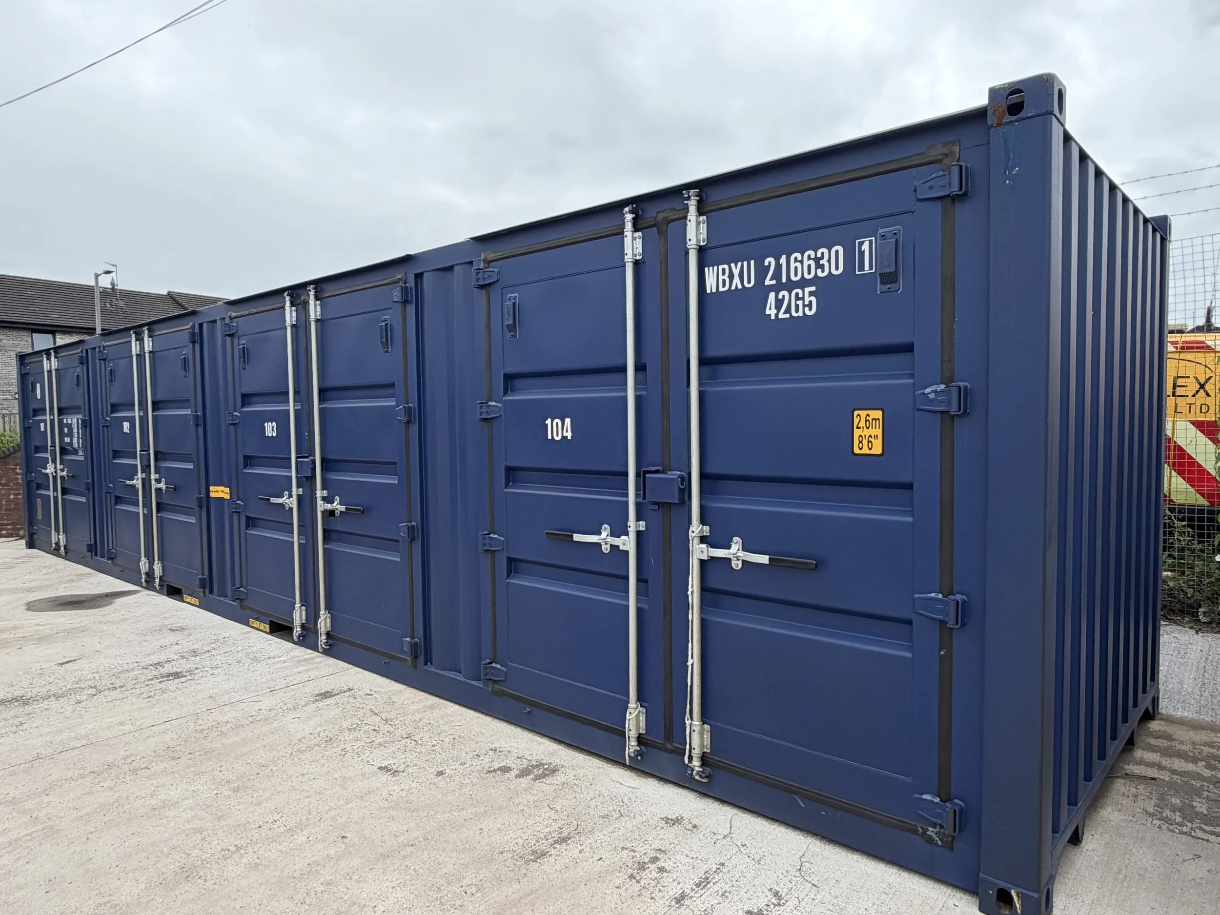 Blue shipping container with white identification numbers and locking mechanisms, situated outdoors on a concrete surface with a residential building and cloudy sky in the background.