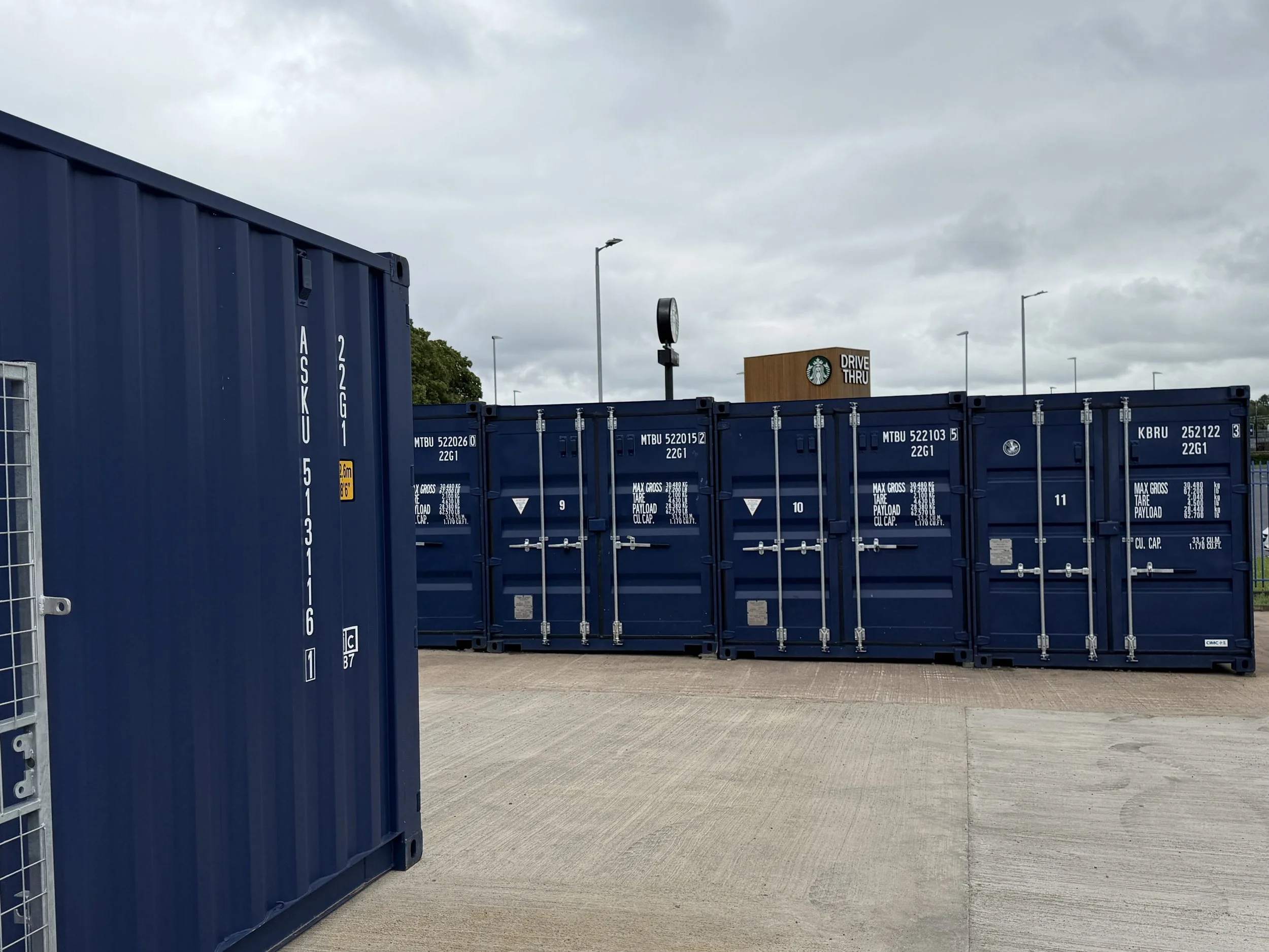 Blue shipping containers in an outdoor lot with a Starbucks drive-thru sign and streetlights under cloudy skies.