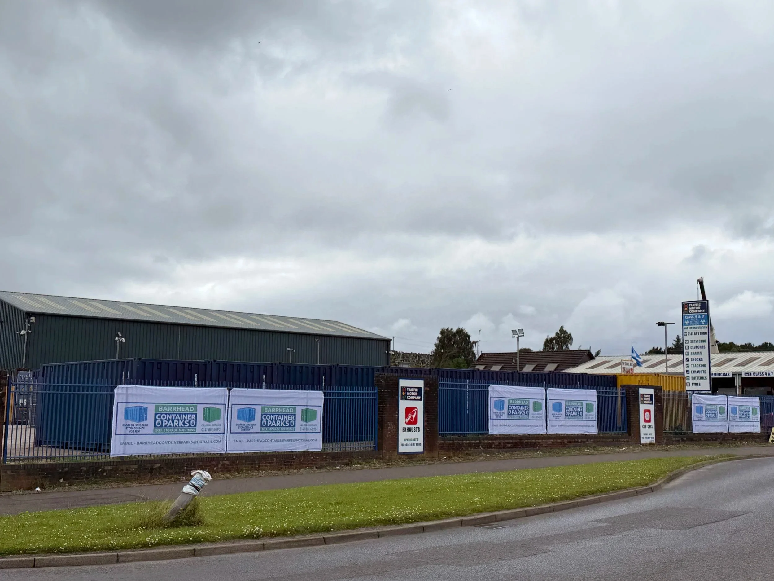 A commercial building with a blue fence and signs advertising container parks and storage solutions. Overcast sky with dark clouds and some trees in the background.