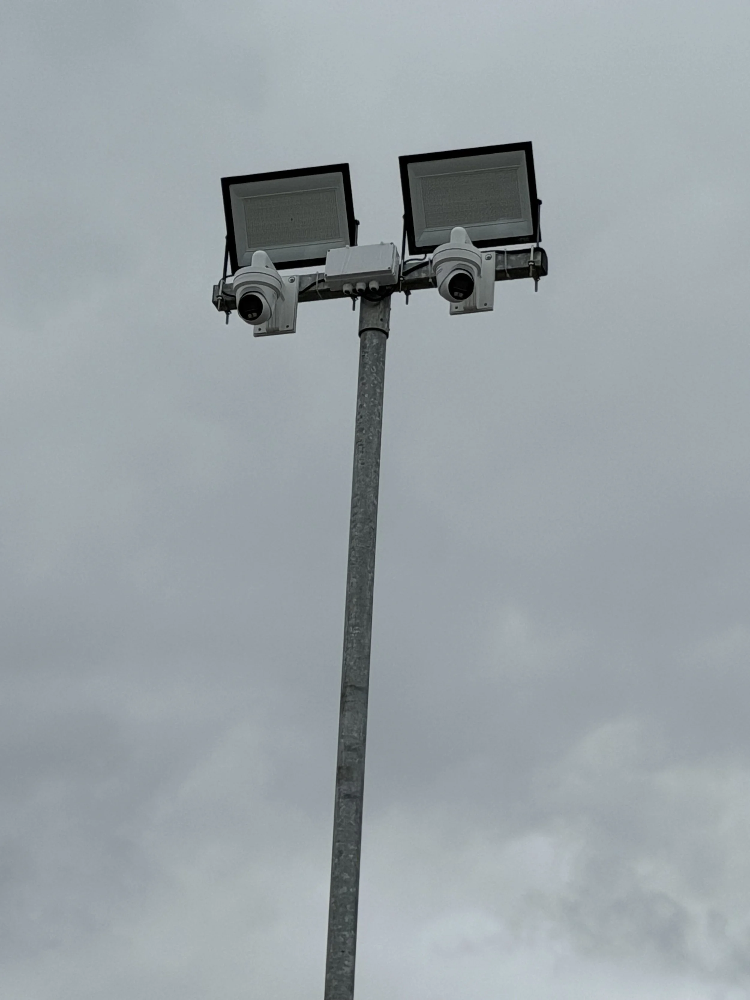 A tall streetlight pole with two floodlights and security cameras attached, set against a cloudy sky.