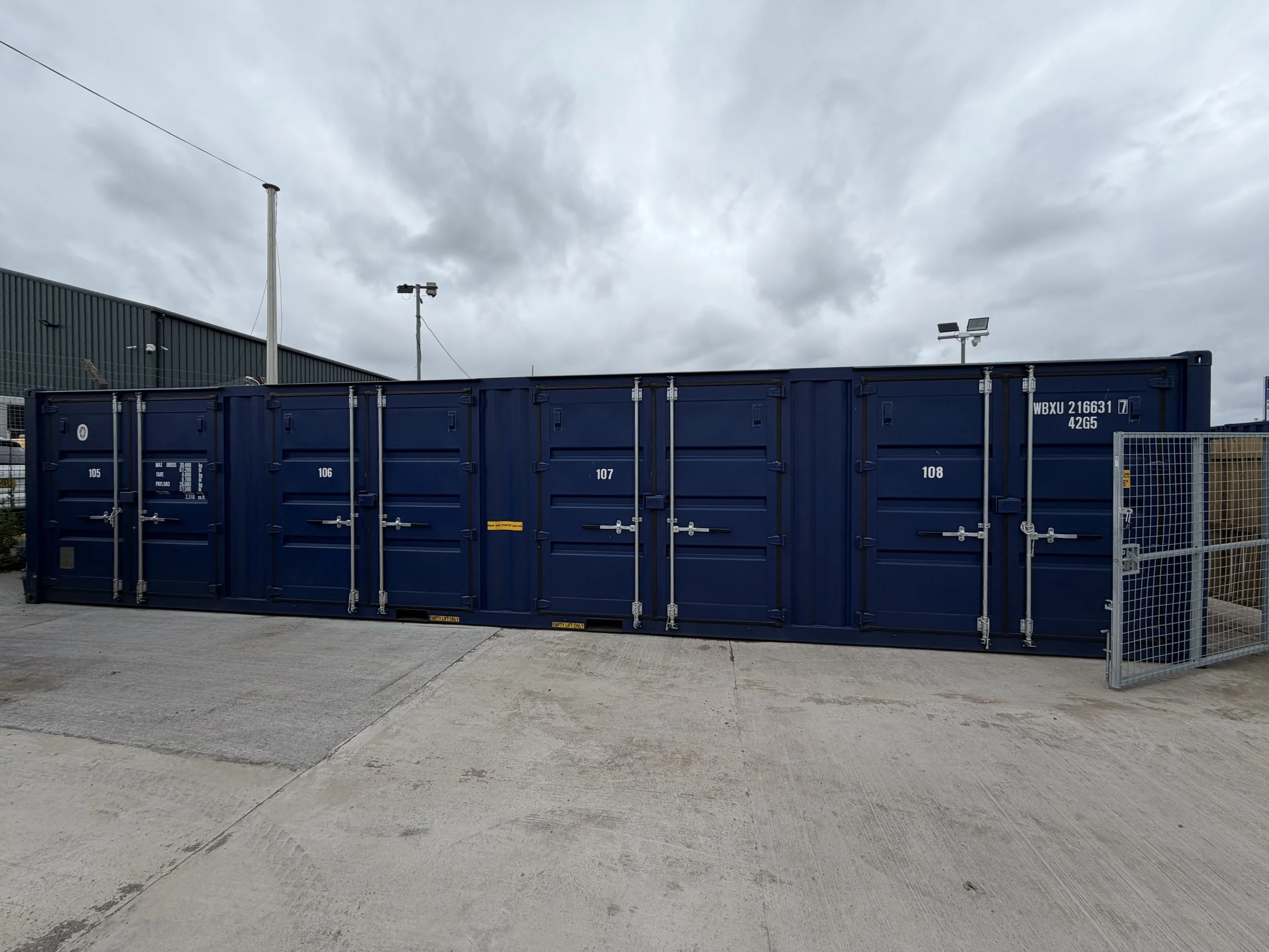 View of a row of blue shipping containers on a concrete lot under cloudy sky.