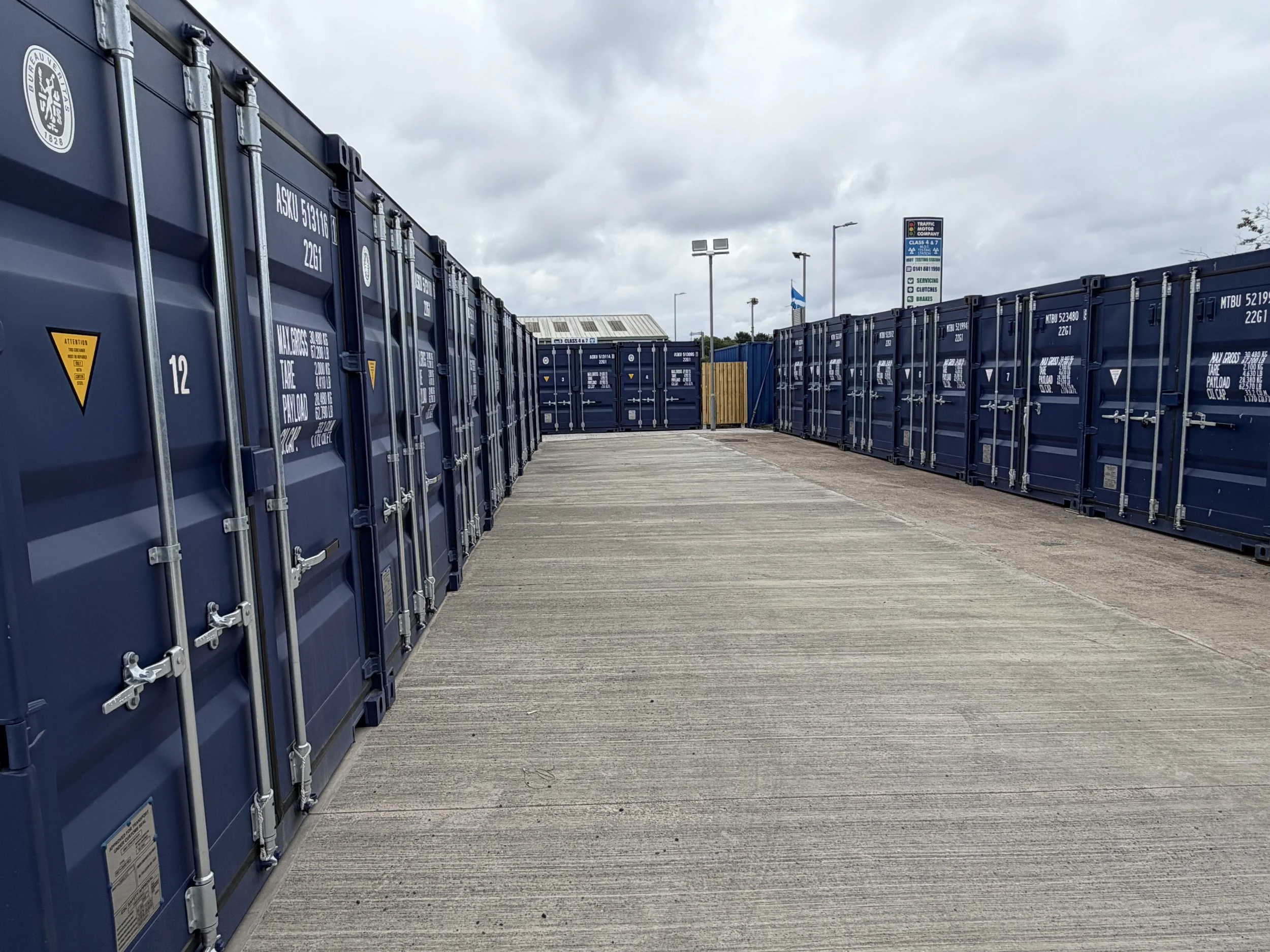 A parking lot filled with blue storage containers and overhead parking lot signs, with cloudy skies above.
