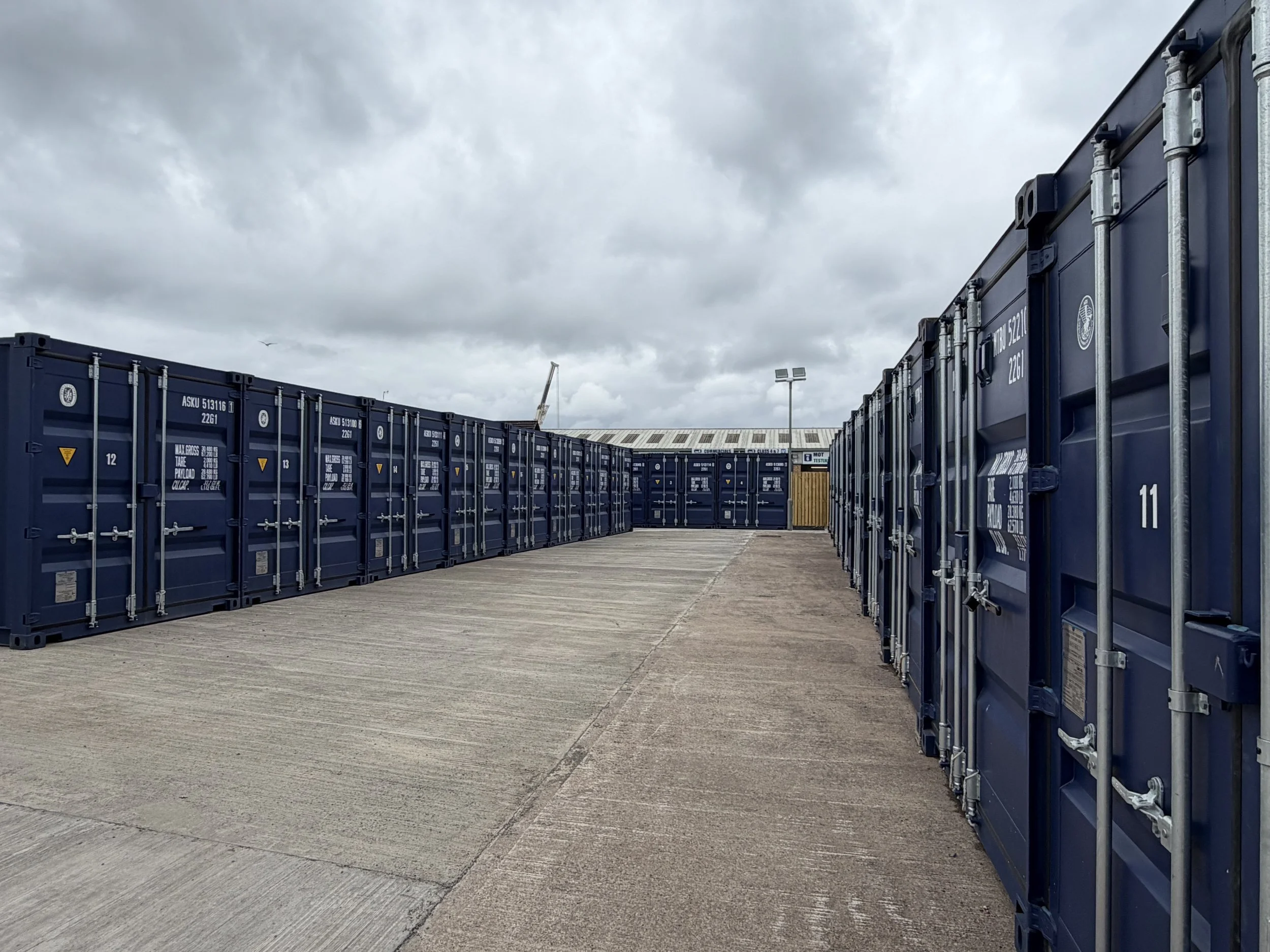 A row of blue shipping containers on a concrete surface at an outdoor storage yard. Overcast sky with dark clouds and a crane in the background.