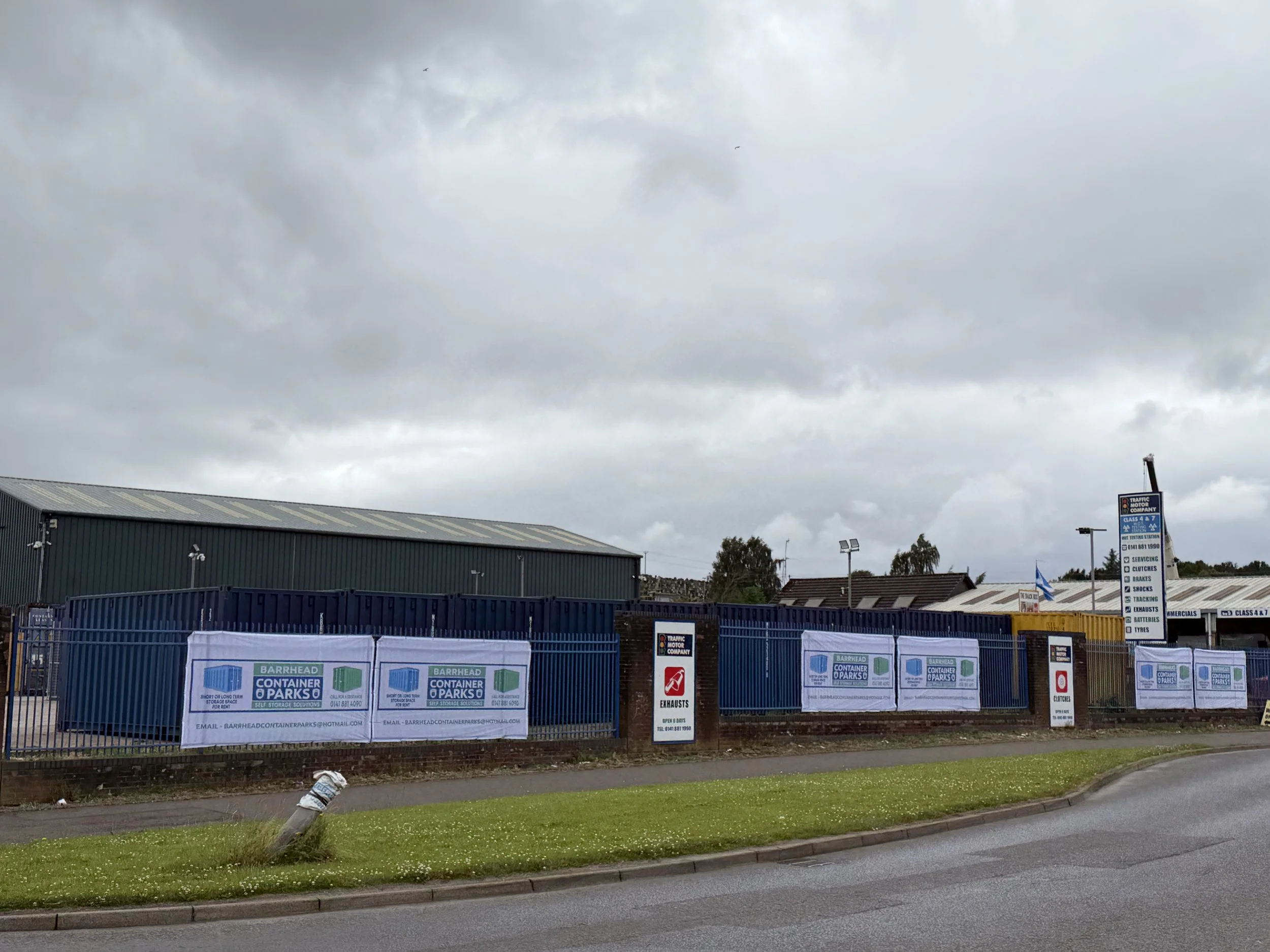 A fence decorated with banners that have the logo for Barrhead Container Parks, an industrial or commercial building with a sign about services like exhausts and shocks, and a cloudy sky overhead.