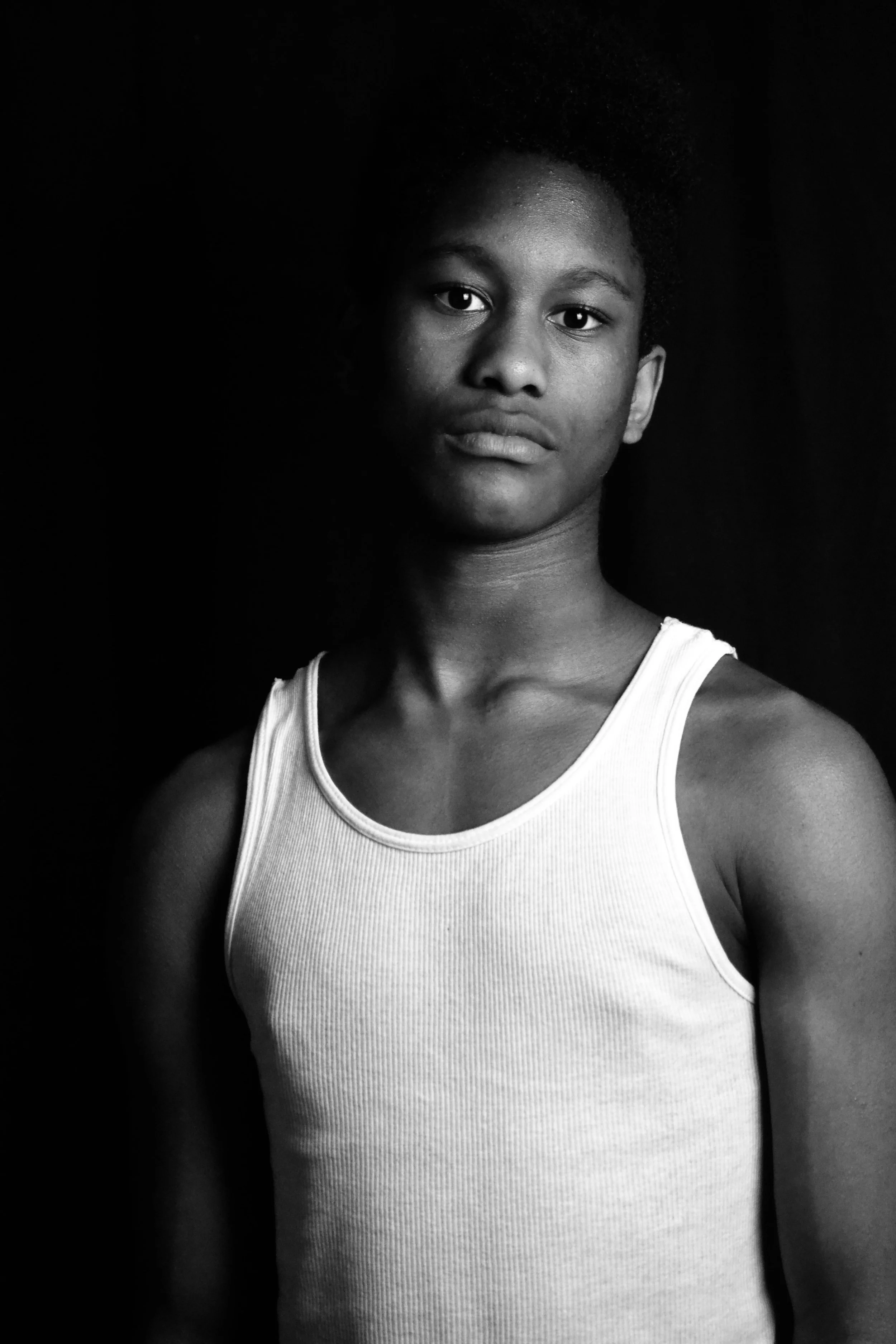 Black and white portrait of a woman with short curly hair wearing a tank top, looking directly at the camera with a neutral expression, against a dark background.