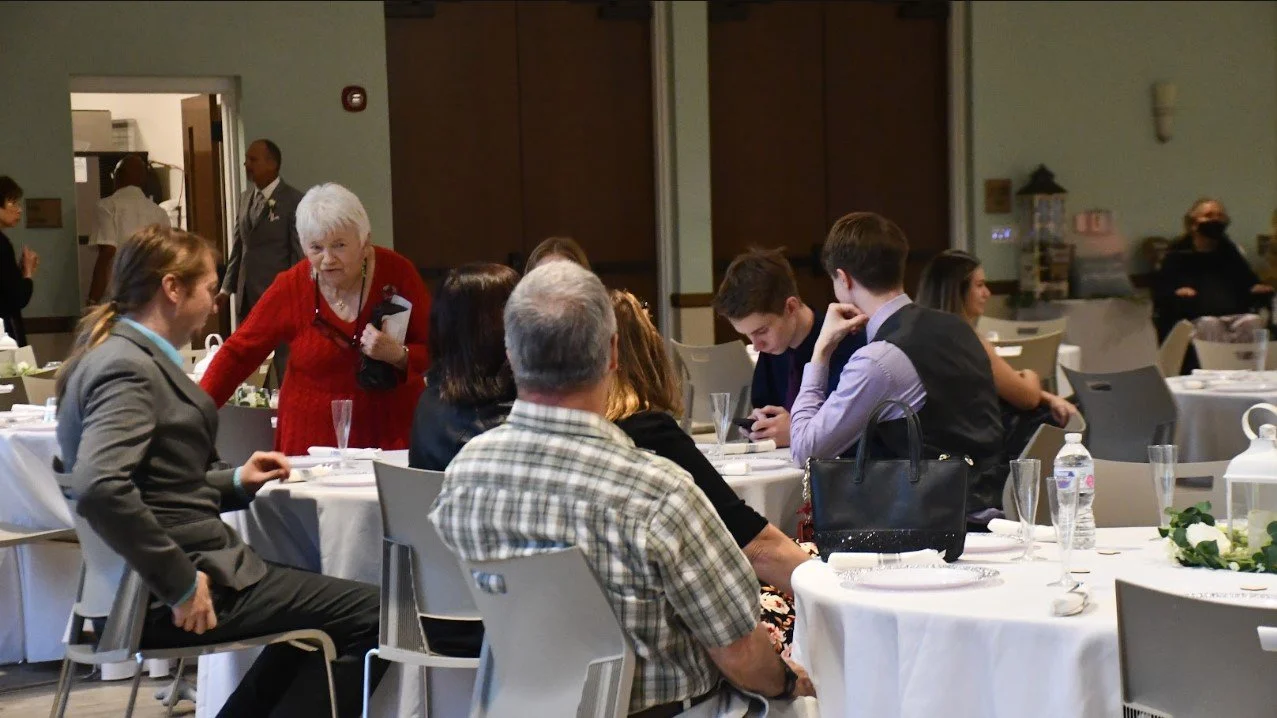 Group of people seated at several round tables during a social event in a banquet hall, with some individuals engaged in conversation and others looking at their phones.