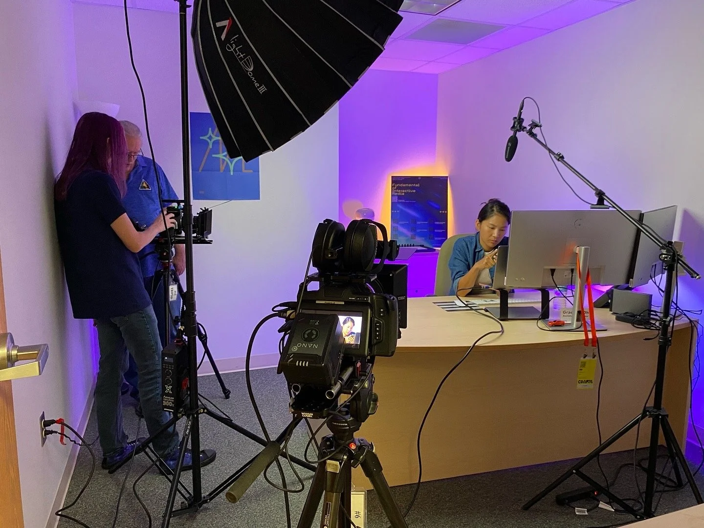 A woman sitting at a desk with two computer monitors, working on a computer, with professional video filming equipment and crew members filming her in a well-lit, small office or studio space.