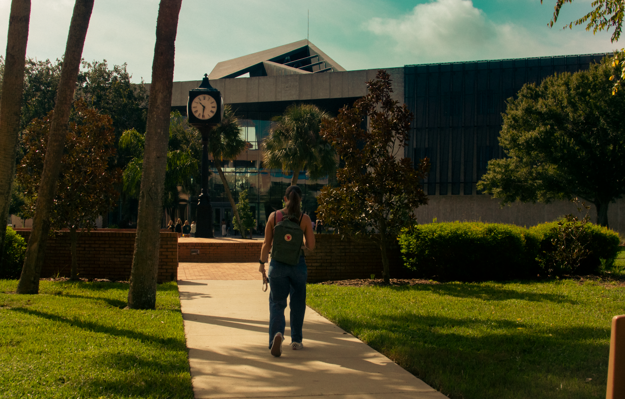 A person with a backpack walking on a sidewalk toward a modern building with a clock tower in front, surrounded by trees and greenery on a sunny day.