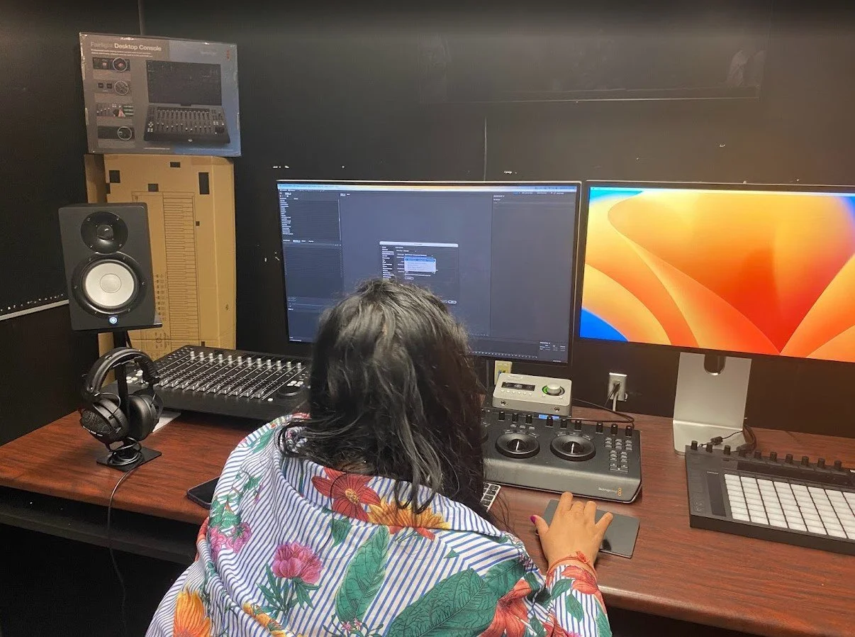 Person with dark hair wearing a floral shirt working at a music production desk with two monitors, a sound mixer, headphones, and a keyboard.