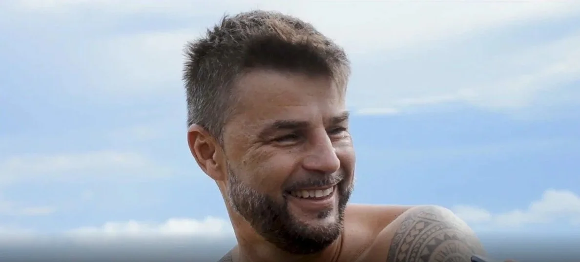 A man with a beard and tattoo smiling outdoors against a cloudy sky