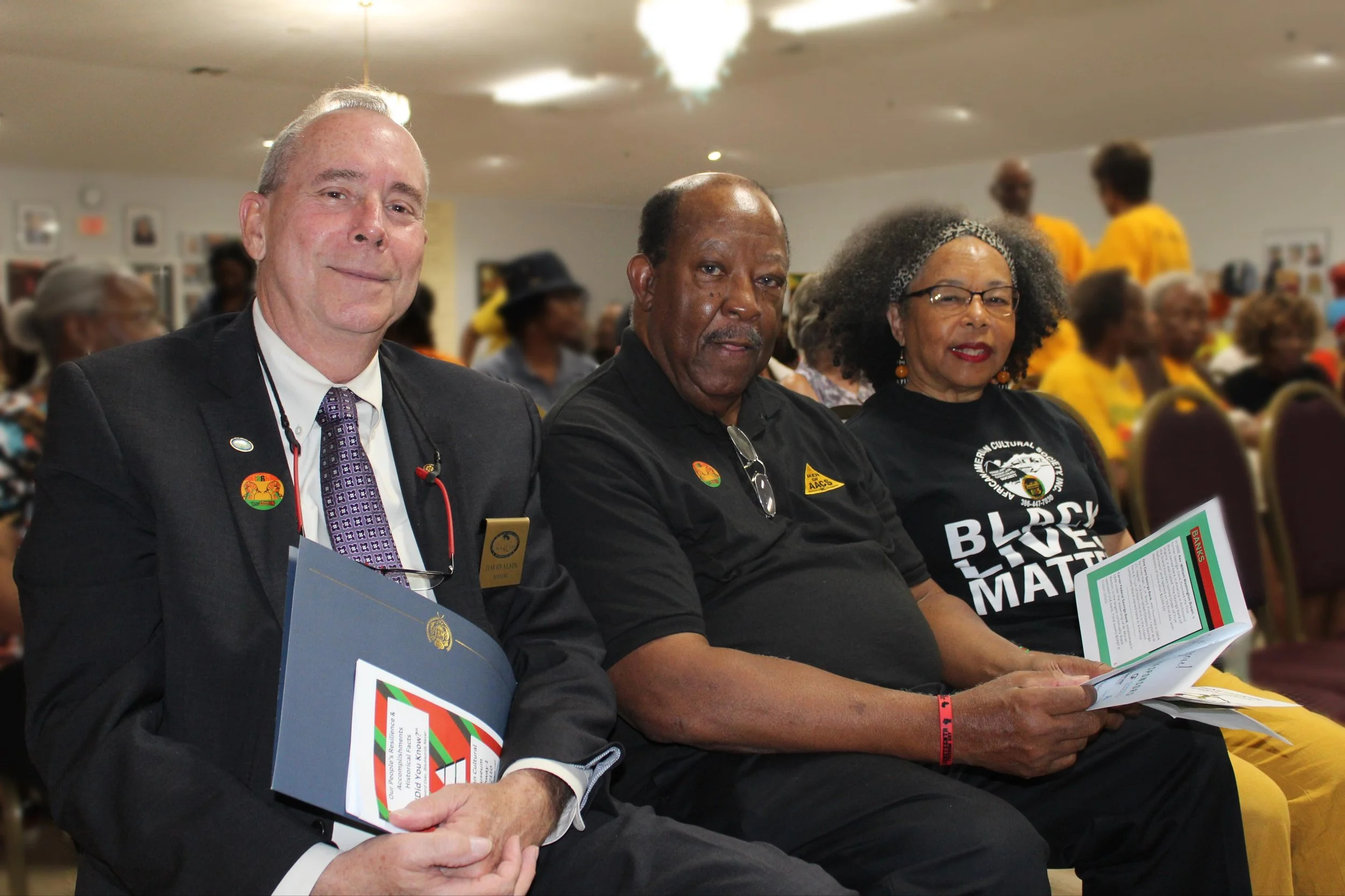 Three adults sitting in a conference room, holding pamphlets, with other people seated in the background, some wearing yellow shirts. The man on the left is dressed in a suit with a tie and has a badge, the man in the middle has a black shirt with a sticker, and the woman on the right wears glasses and a black t-shirt with bold white text.