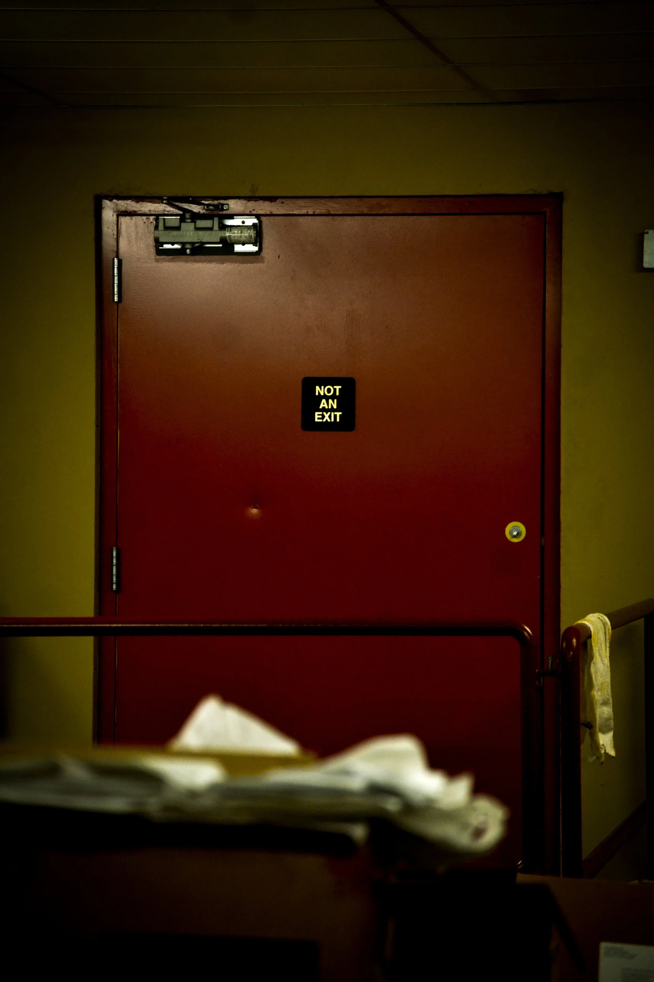 Red door with a sign that says 'NOT AN EXIT' in illuminated yellow letters, keypad and door closer at the top, beige walls, some papers on a table in the foreground, towel hanging on the right side.