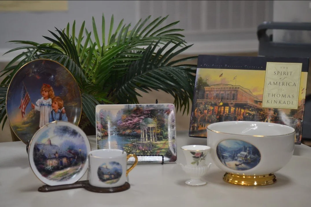 Collection of decorative items and memorabilia on a table, including plates, a teacup, a bowl with a gold base, a small cup, framed pictures of American scenes and landmarks, books, and a green plant in the background.