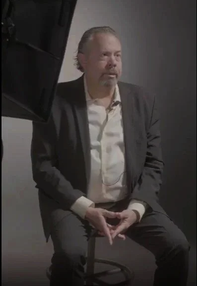 A man with gray hair and a beard, wearing a dark blazer and white shirt, sitting on a stool against a plain dark background, looking to the side with a serious expression.