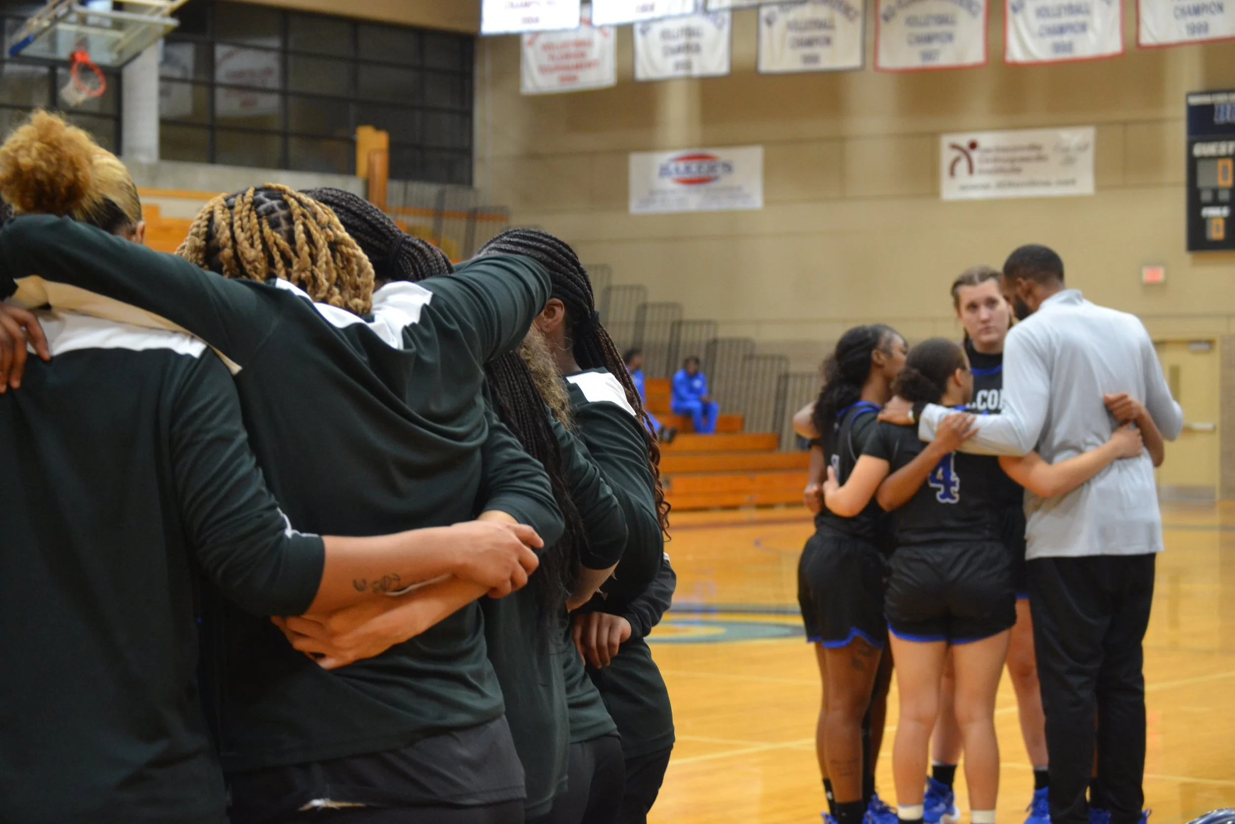A group of female basketball players and their coach standing together in a gym, with their arms around each other in a huddle, during a team bonding moment.