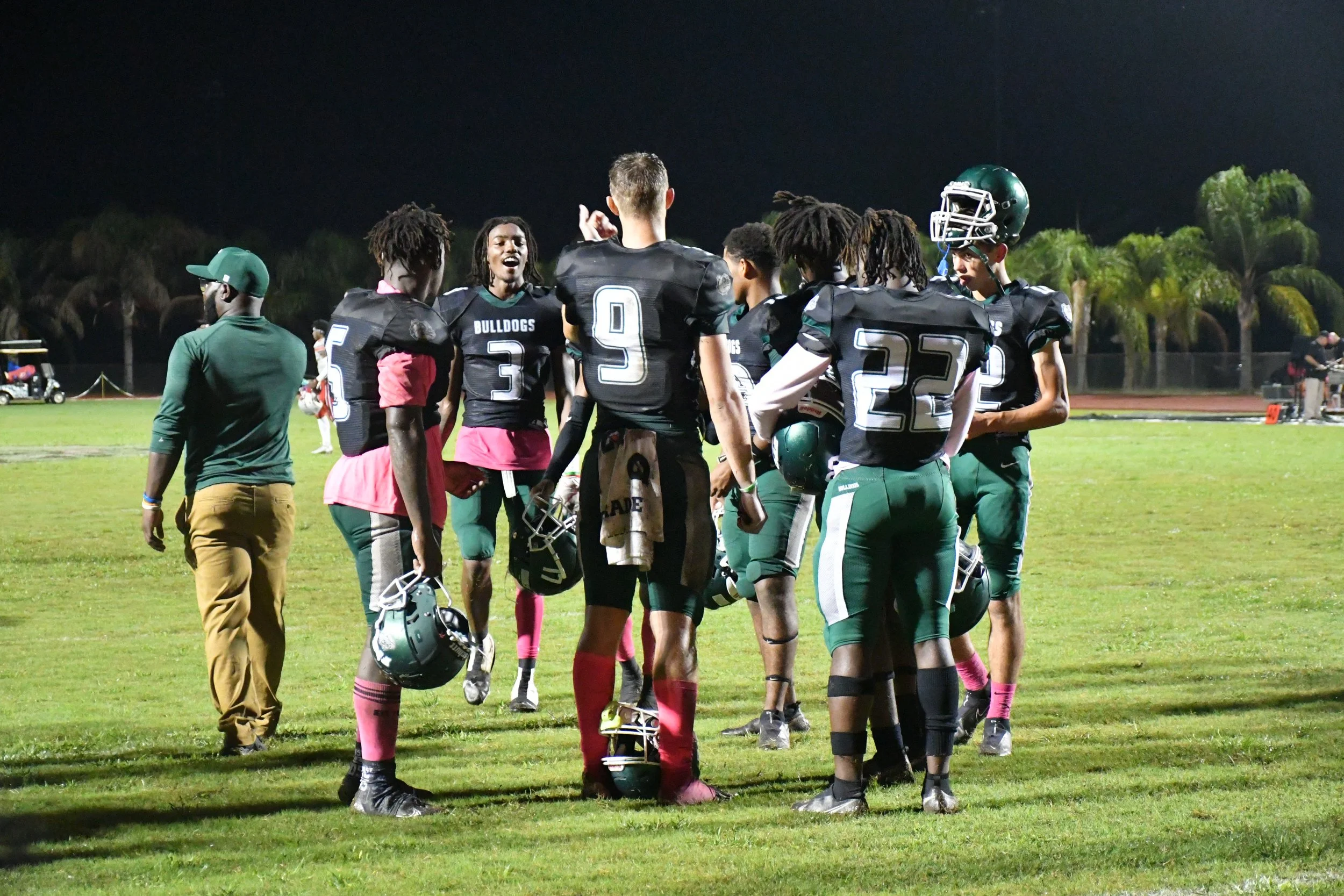 A group of football players wearing black and green uniforms gathered in a huddle on the field during a nighttime game, with some wearing helmets and holding their helmets in their hands.