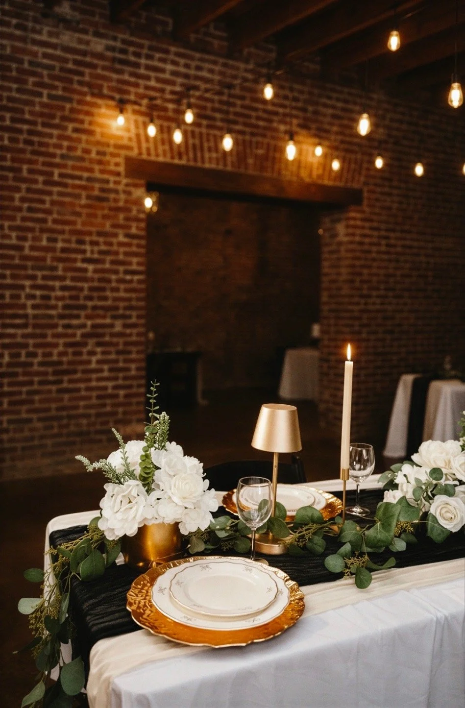 Elegant birthday table setup with floral centerpiece, gold chargers, white and gold plates, candles, and greenery, in a rustic brick-walled venue with hanging string lights.