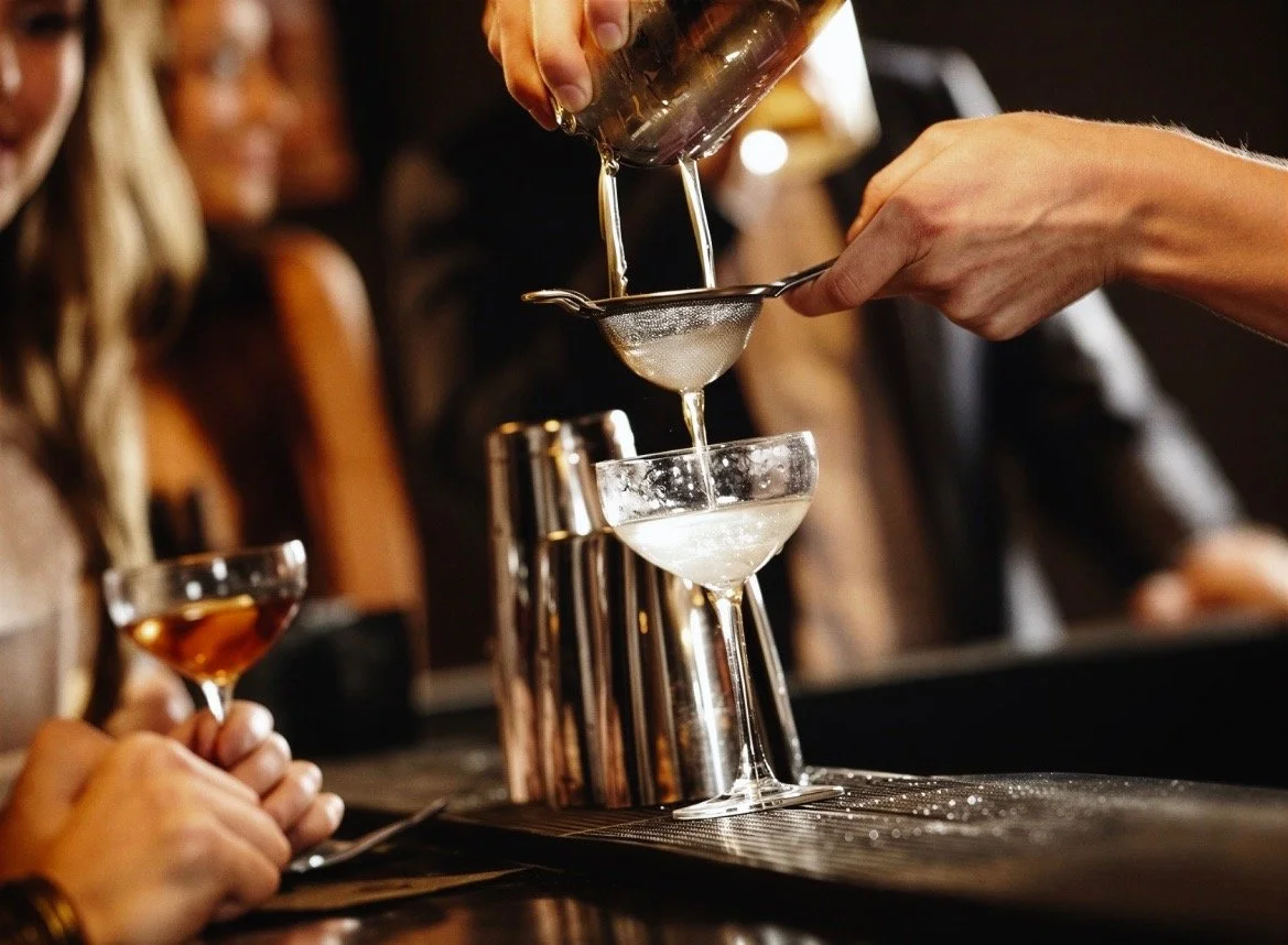 Bartender pouring a cocktail through a strainer into a coupe glass at a bar, with a group of women watching.