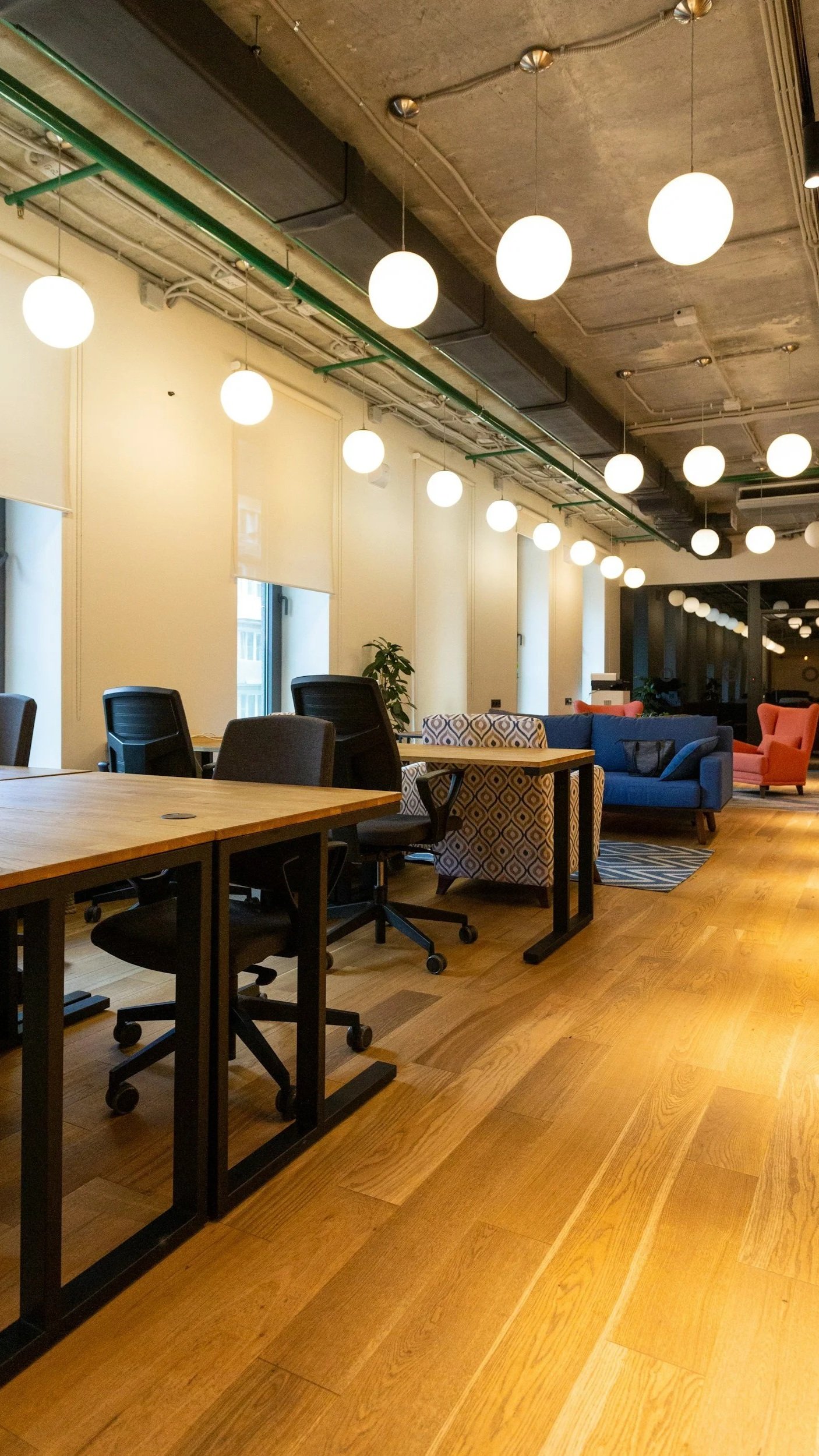 Modern office lounge with wooden floors, black office chairs, tables, a blue sofa, orange armchair, patterned armchair, and large windows; ceiling has exposed concrete with round globe pendant lights.