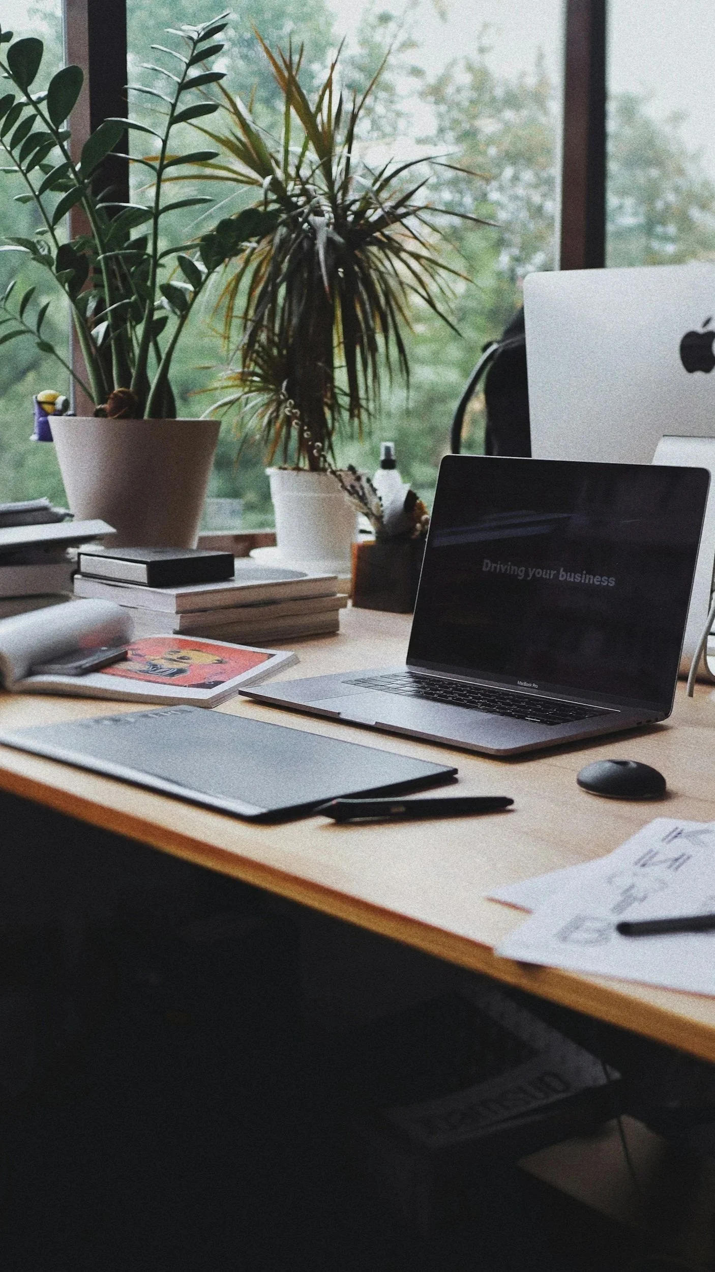 A tidy office desk with a laptop displaying the phrase 'Driving your business,' a desktop computer, books, stationery, and potted plants next to a large window showing trees outside.