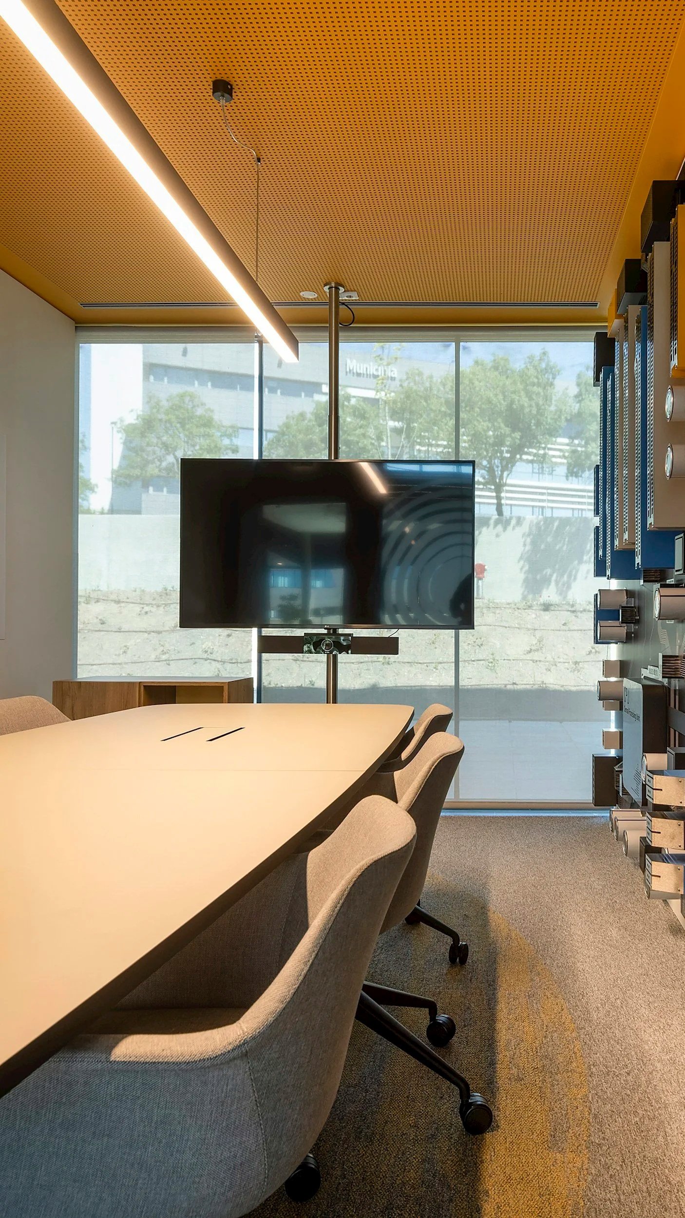 A modern conference room with a rectangular table surrounded by beige chairs on wheels, a large flat screen TV on a stand, a ceiling with wooden panels and integrated lighting, and a large window showing trees and a building outside.
