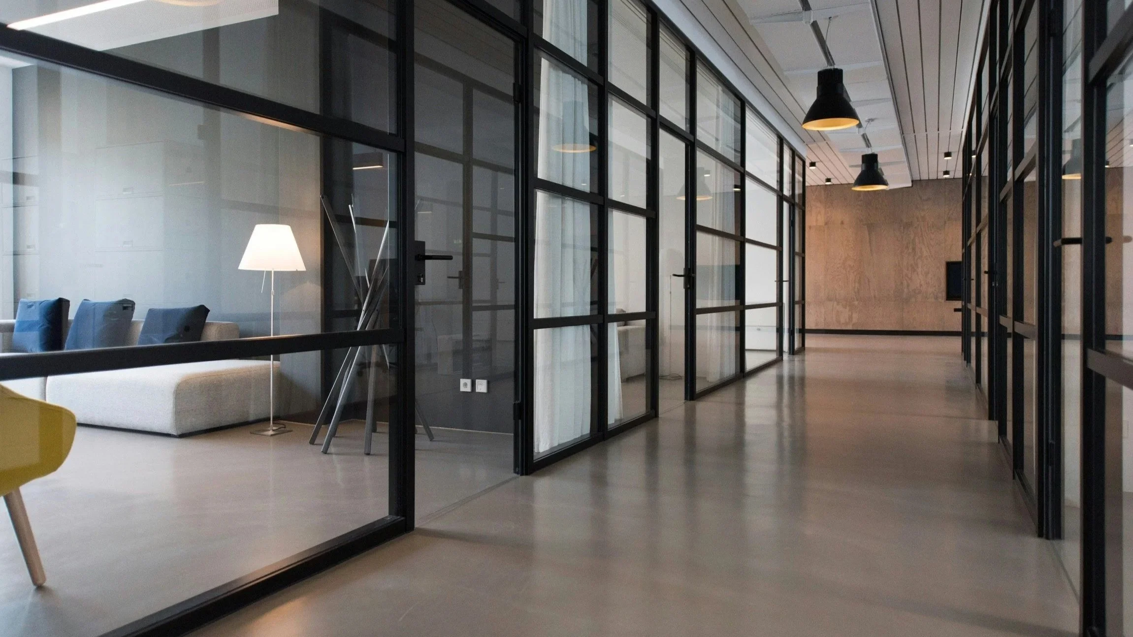 Modern office space with glass partitions, a seating area with a lamp, and contemporary lighting fixtures.