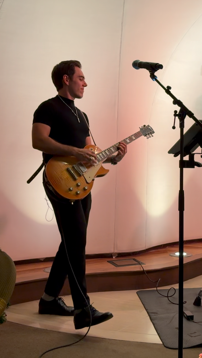 A young man playing an electric guitar on a small stage with a microphone stand nearby.