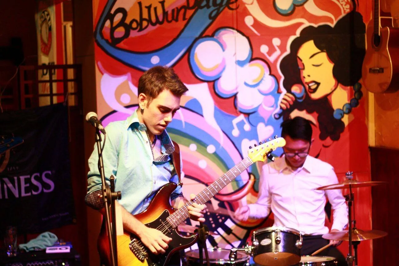 Two young men performing music on stage with a colorful cartoon mural of a woman singing in the background.