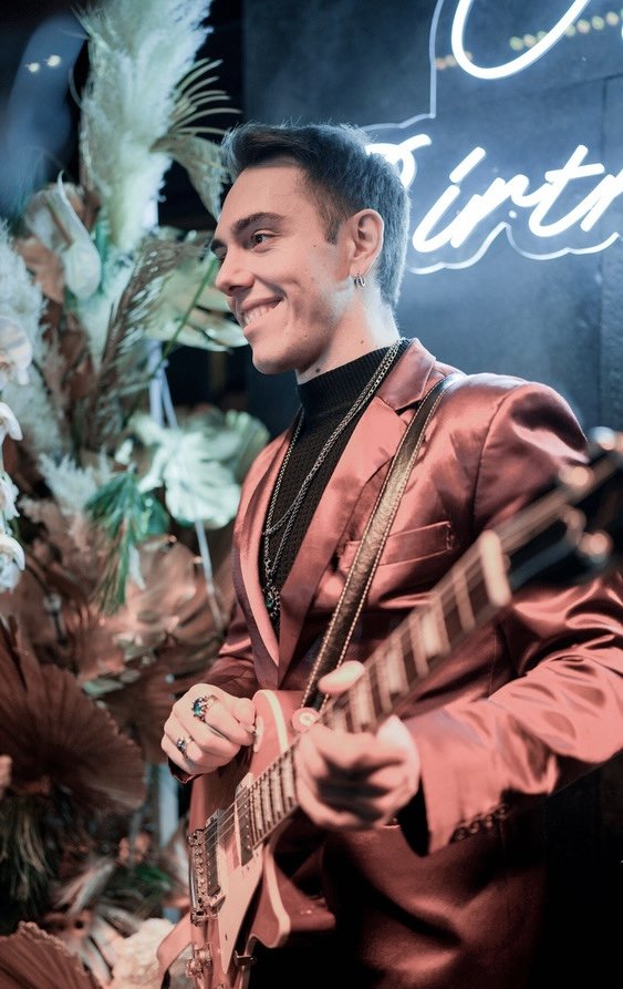 A young man playing an electric guitar at a party, smiling, wearing a pink satin blazer and black shirt, in front of a decorative backdrop with dried flowers and a neon sign.