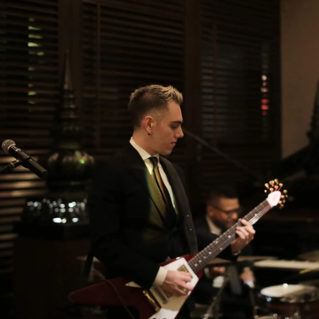 Young man with short, styled hair, dressed in a black suit, playing an electric guitar in a dimly lit room with wooden blinds. A man in the background is sitting at a piano, smiling.