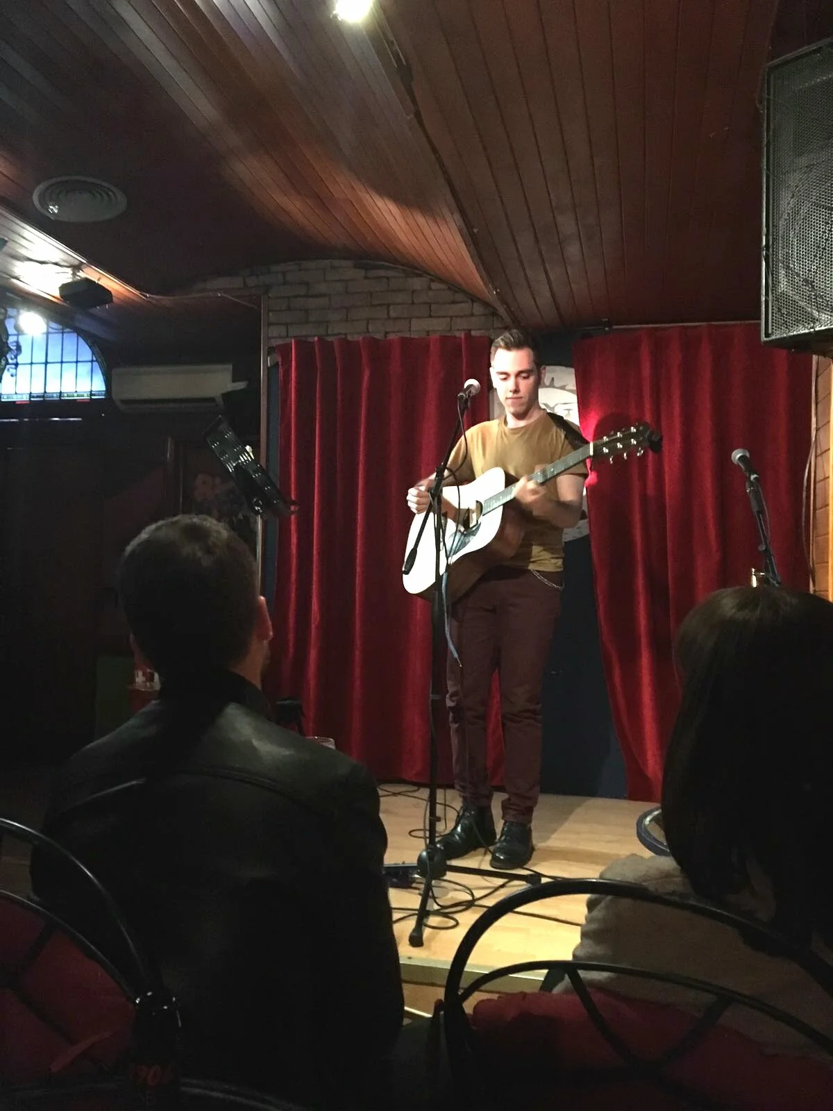 A young man performs on stage with an acoustic guitar, standing in front of red velvet curtains inside a bar or club. Audience members are watching him, some visible from behind.