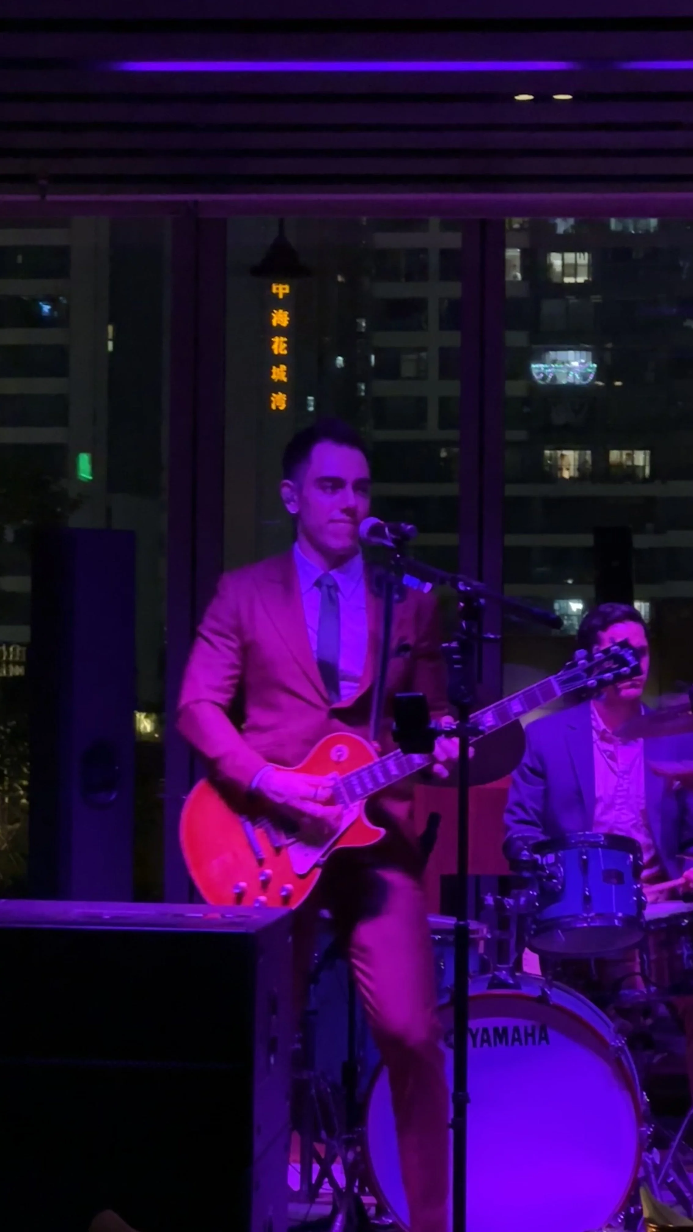A musician playing an orange electric guitar and singing into a microphone during a performance, with a city skyline and illuminated signs visible in the background.