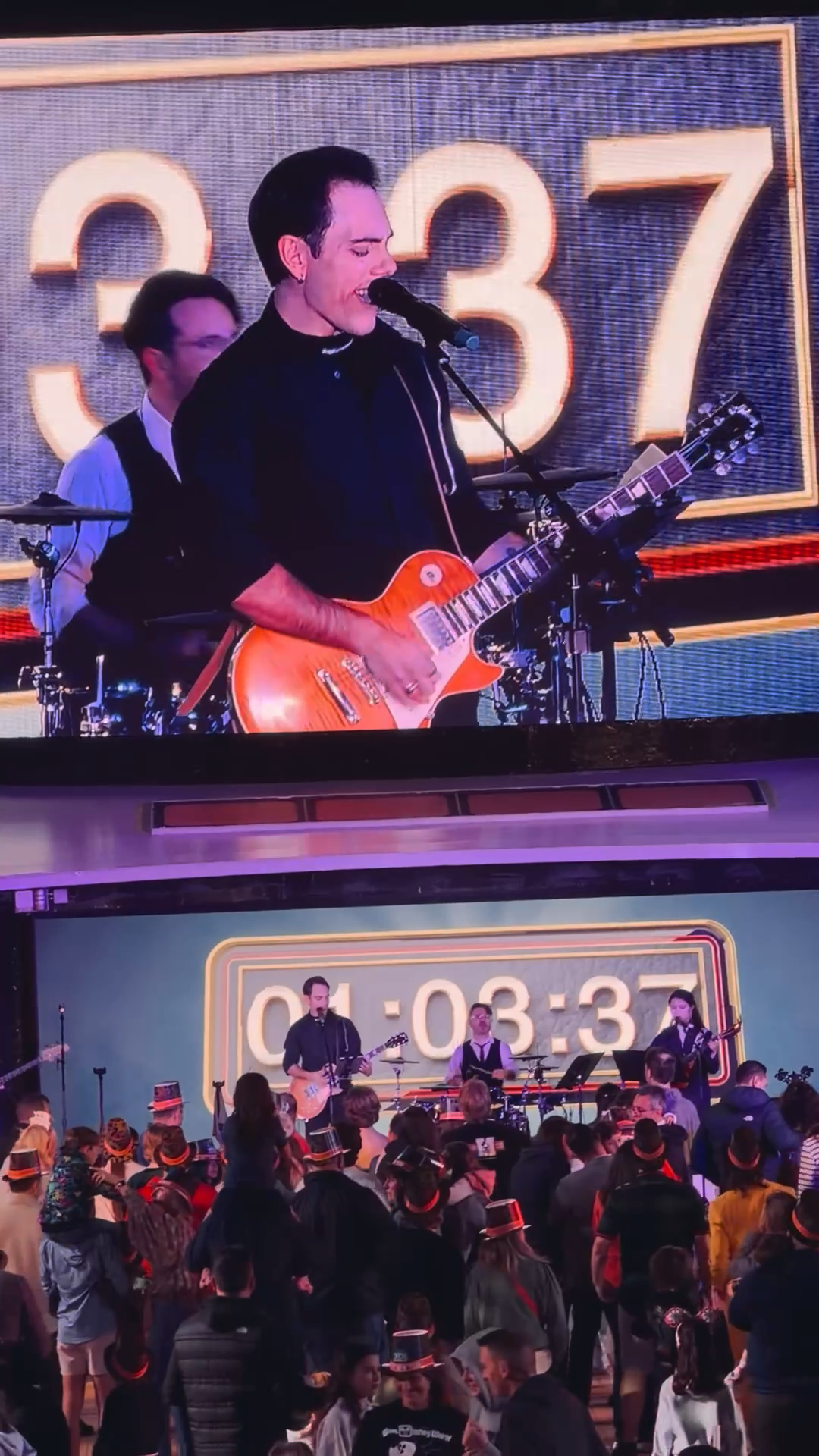 A band performs on stage with a large timer in the background showing 3 minutes and 37 seconds. The lead singer is playing an orange electric guitar and singing into a microphone. There is an audience watching the performance.