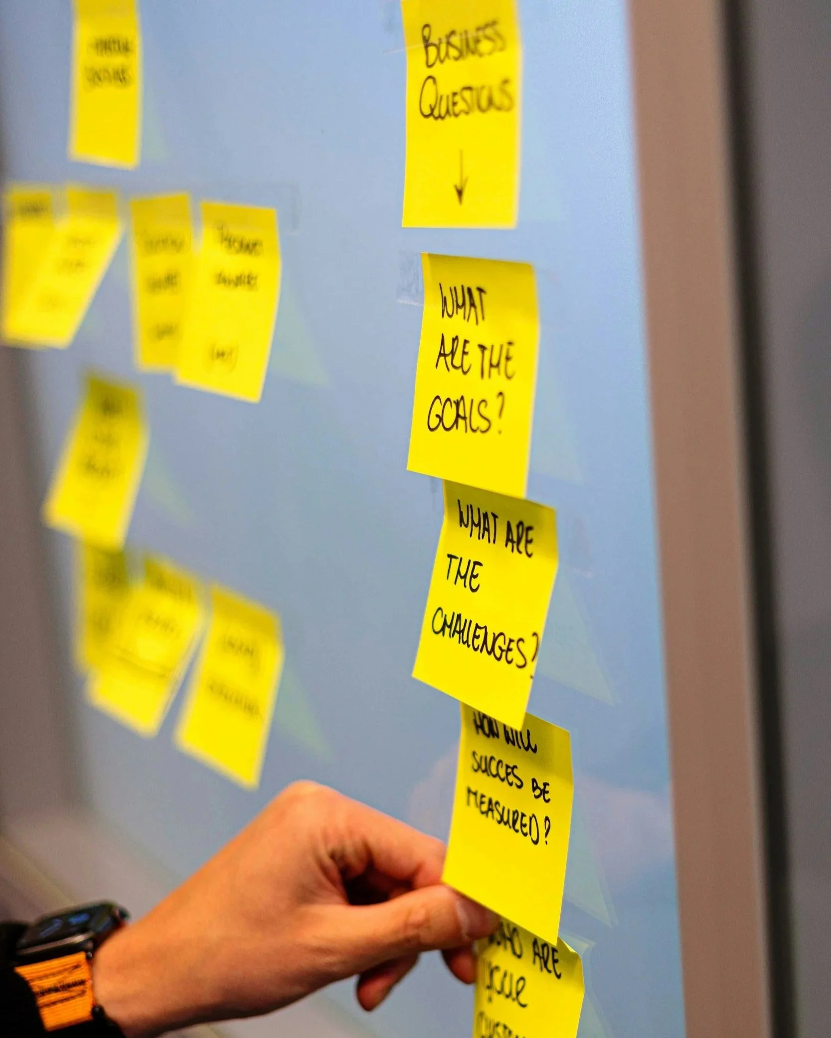 A person holding a yellow sticky note with the handwritten question, 'What are the challenges?' It appears to be part of a larger collection of notes on a wall, with other sticky notes visible in the background. The person is wearing a black and orange striped wristwatch.