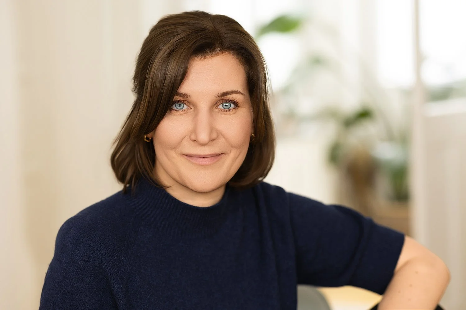 Eine Frau mit braunen, schulterlangen Haaren und blauen Augen, trägt ein dunkelblaues Oberteil und lächelt leicht in einem hellen Raum mit Pflanzen im Hintergrund.