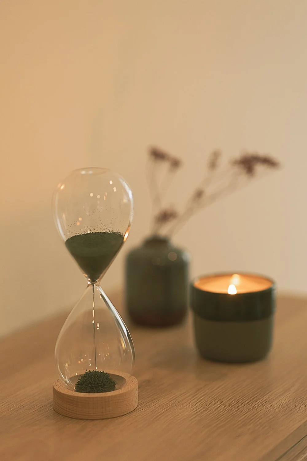 Sanduhr, Vase mit Trockenblumen und Kerze auf einem Holztisch in warmem Licht.