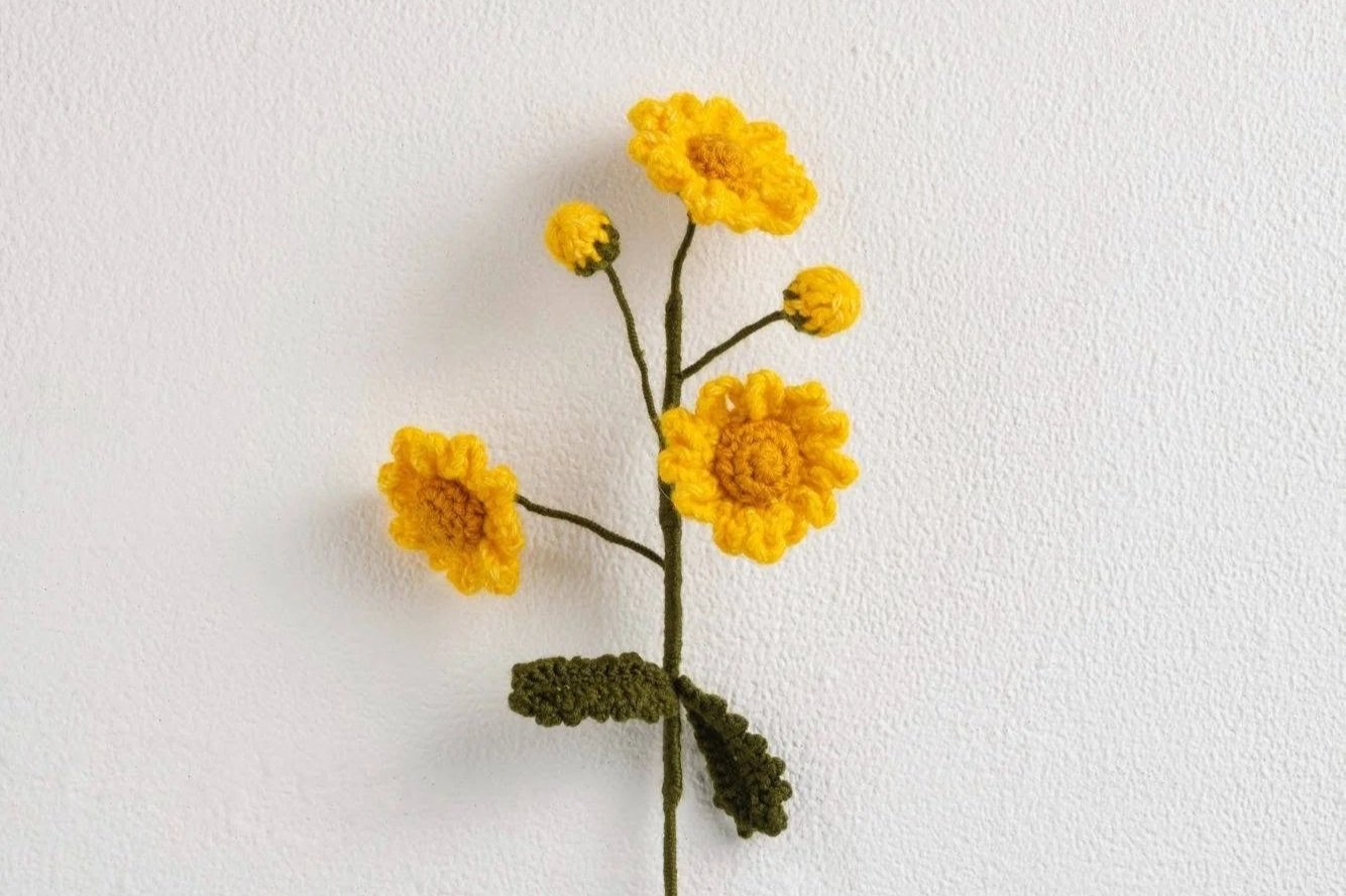 Yarn-crafted yellow flowers with green leaves on a textured white background.