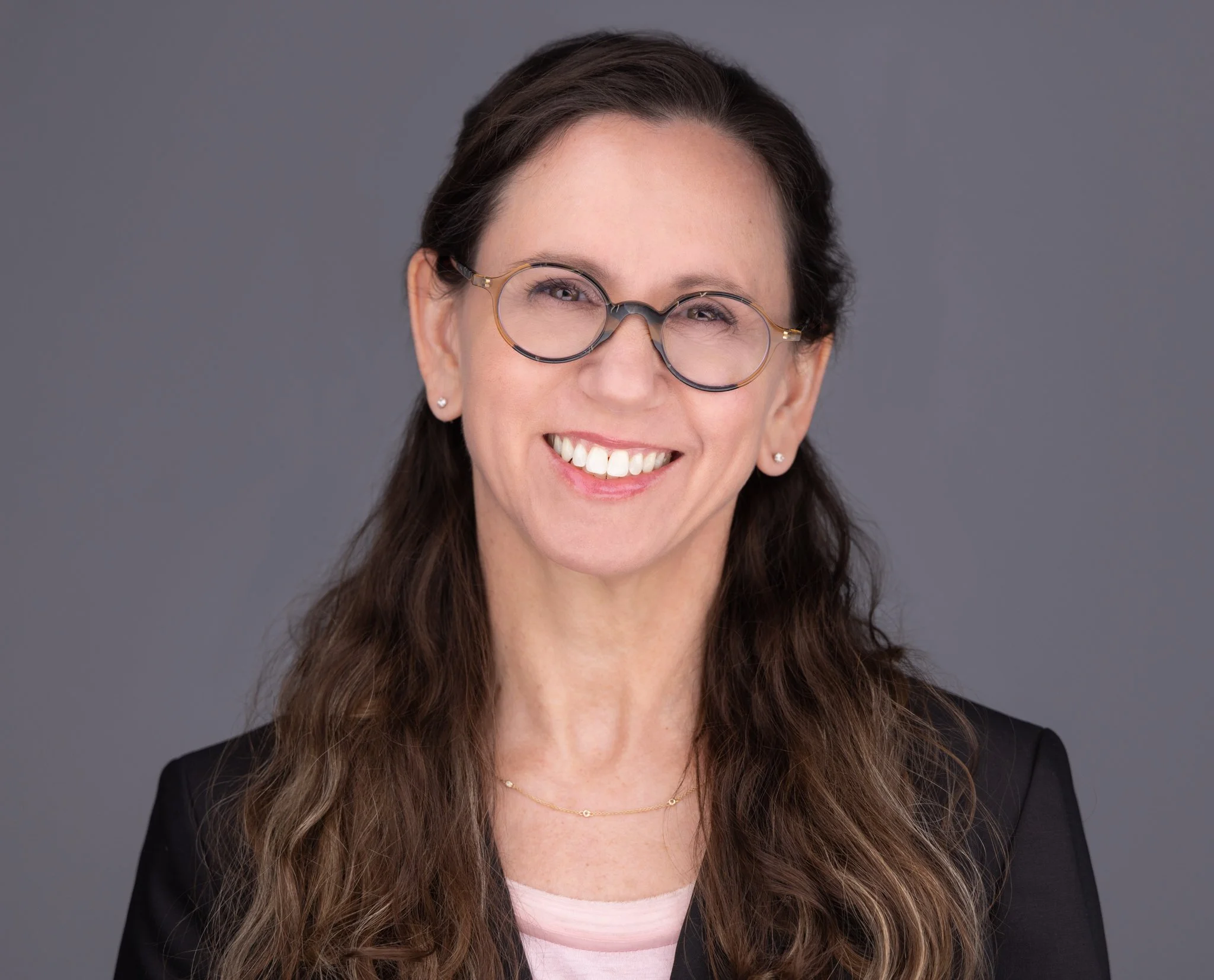 A woman with long curly hair wearing glasses, earrings, a pink top, and a black blazer, smiling against a gray background.