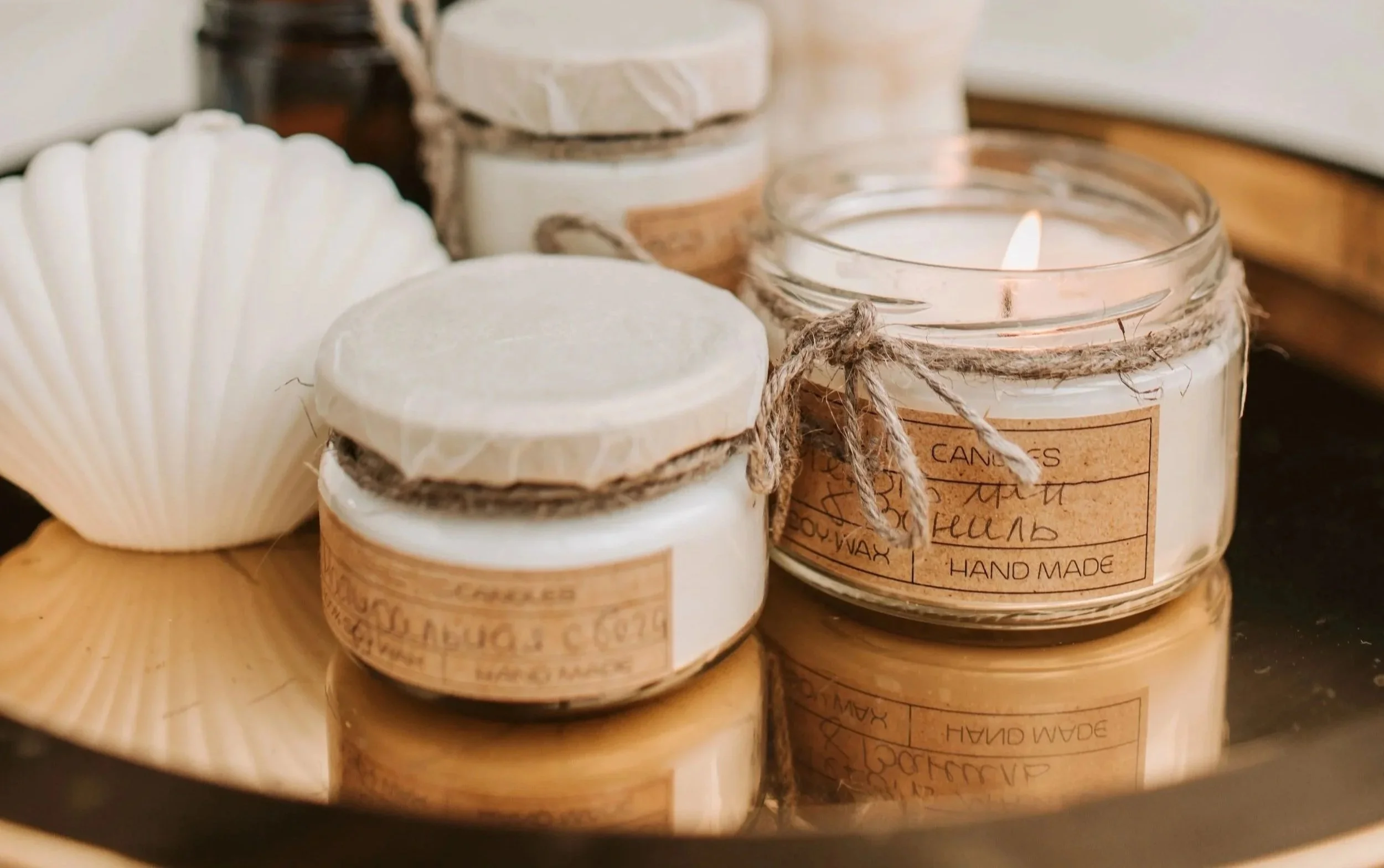 Close-up of several handmade candles in glass jars with twine around the lids, placed on a wooden surface, with one candle lit.