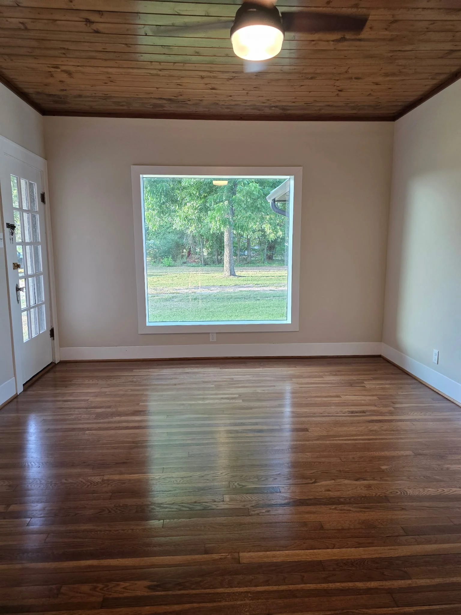 Empty room with hardwood floor, cream-colored walls, a white-framed window showing outside green trees, a white door with glass panes, and a ceiling light.