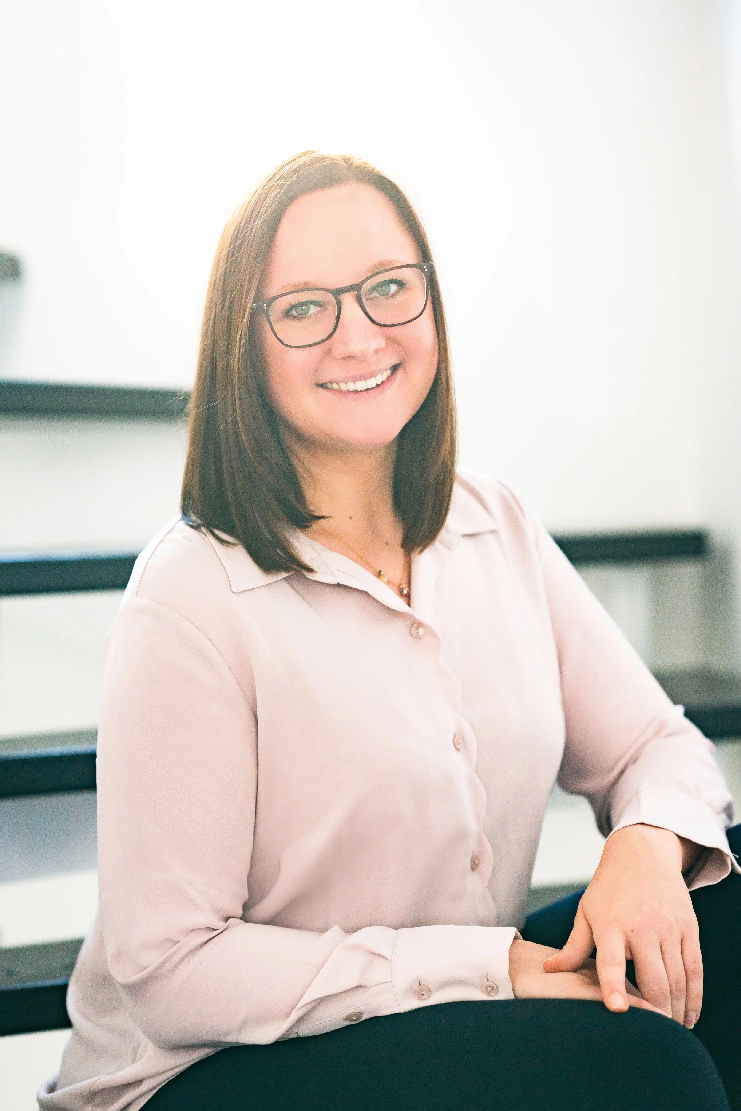 Junge Frau mit Brille lächelt, trägt eine hellrosa Bluse, sitzt an einem Tisch in einem Raum mit hellem Hintergrund.