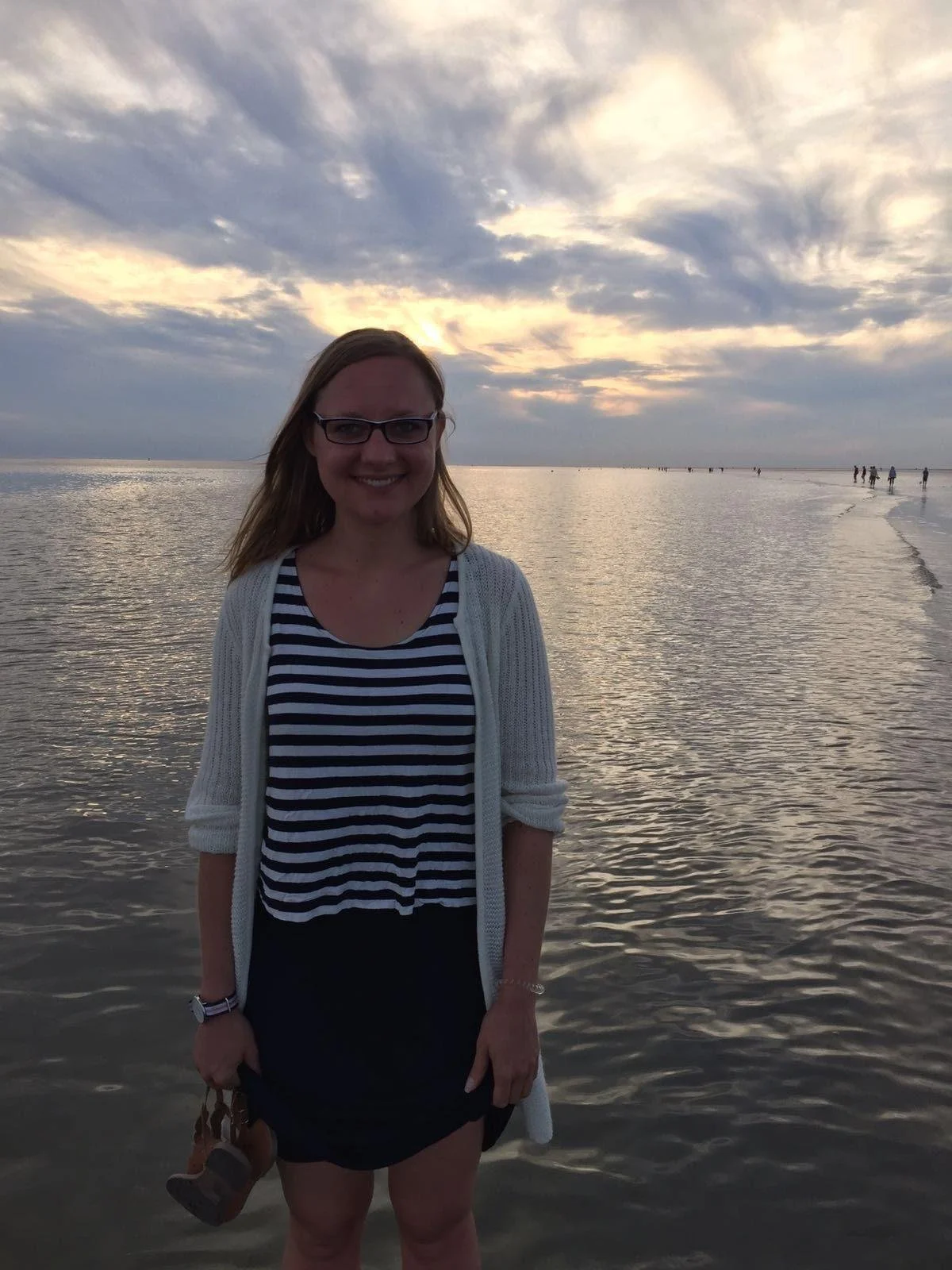 Junge Frau steht am Strand bei Sonnenuntergang, trägt eine schwarze und weiße gestreifte Bluse, einen weißen gestrickten Cardigan und schwarze Shorts, hält Schuhe in der Hand, im Hintergrund sind Wasser, Himmel mit Wolken und einige wenige Menschen am Ufer sichtbar.