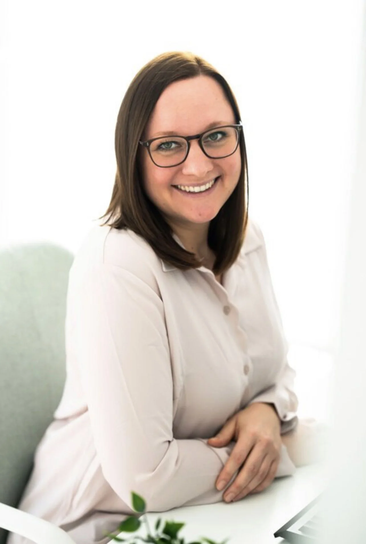 Eine lachende Frau mit Brille sitzt an einem weißen Tisch, trägt eine weiße Bluse und hat braune, glatte Haare.