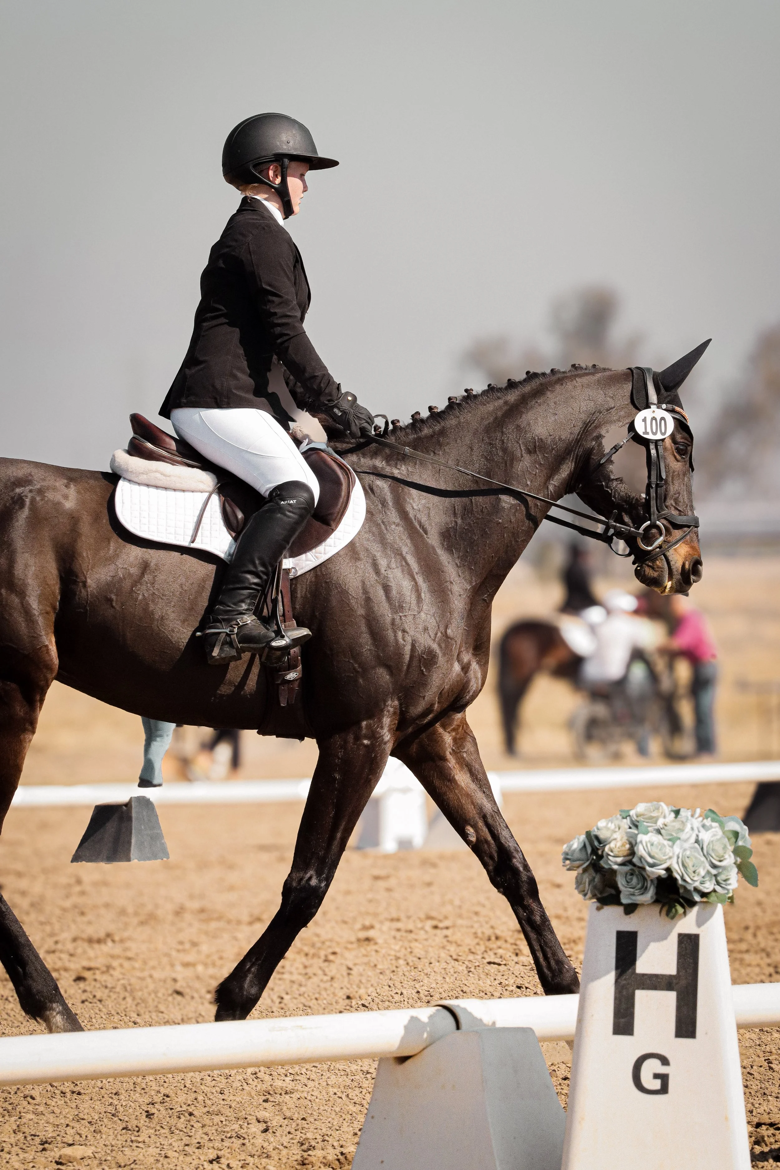 A person riding a brown horse in an equestrian competition, wearing a black helmet, black jacket, white riding pants, and black boots, with a white saddle pad and a white cone with flowers nearby.