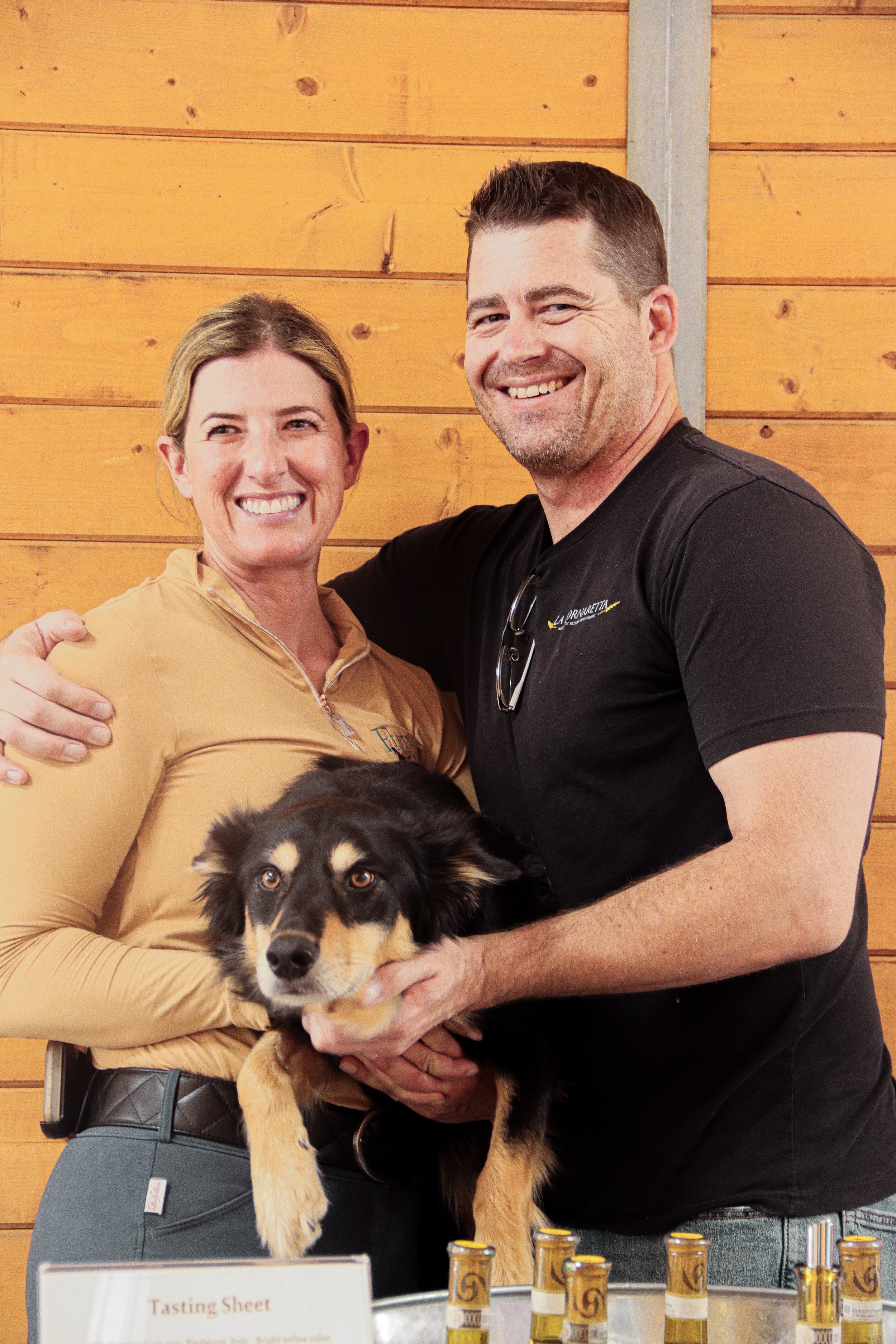 A man and woman smiling and posing together indoors with a wooden wall background, holding a black and tan Australian Shepherd dog. The woman wears a beige zip-up jacket, and the man wears a black T-shirt. There are small bottles on a table in front 