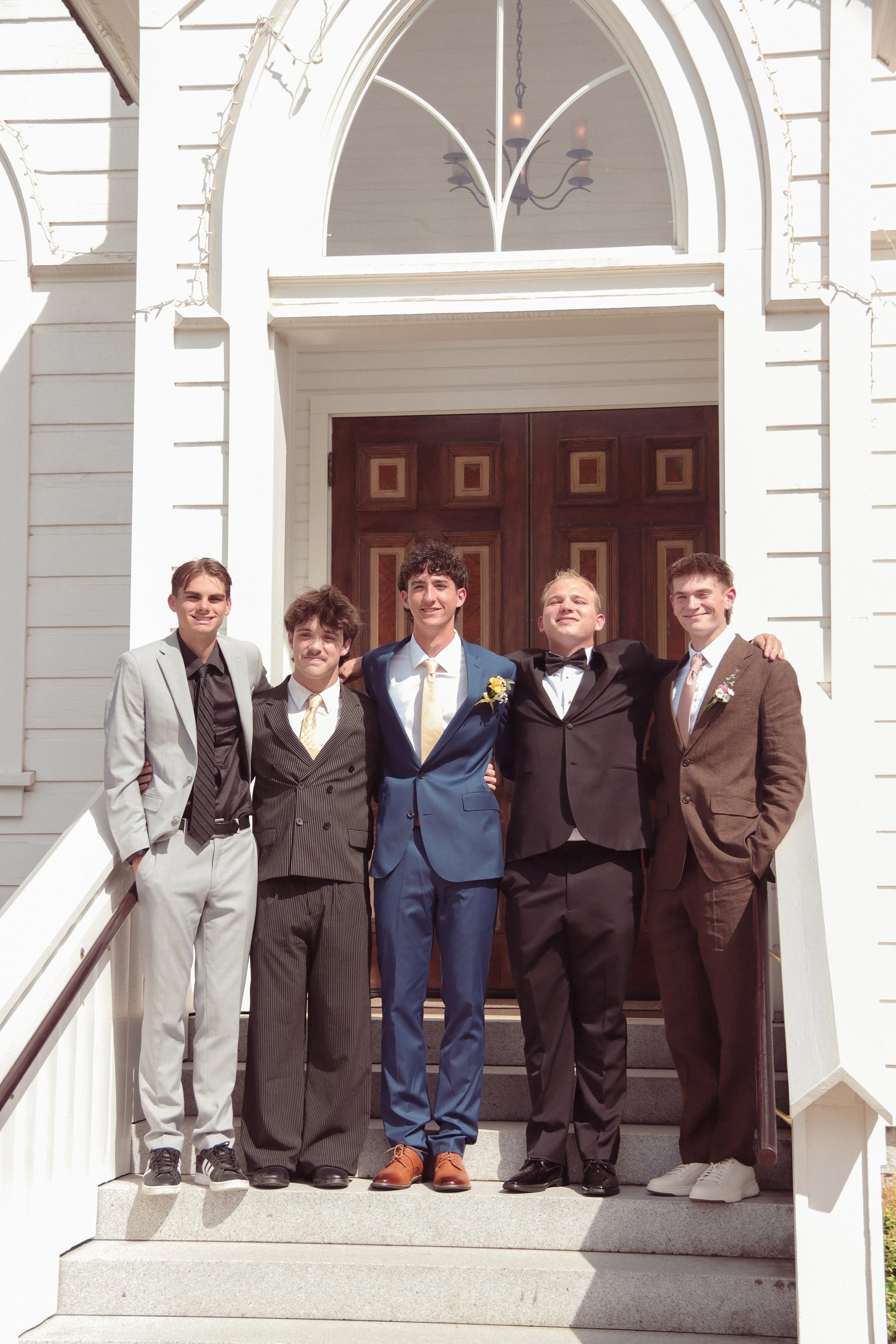 Group of five young men dressed in suits, standing in a line on the steps outside a building, smiling and posing for a photo. The building has a large wooden door, white trim, and a chandelier visible through a window above the door.